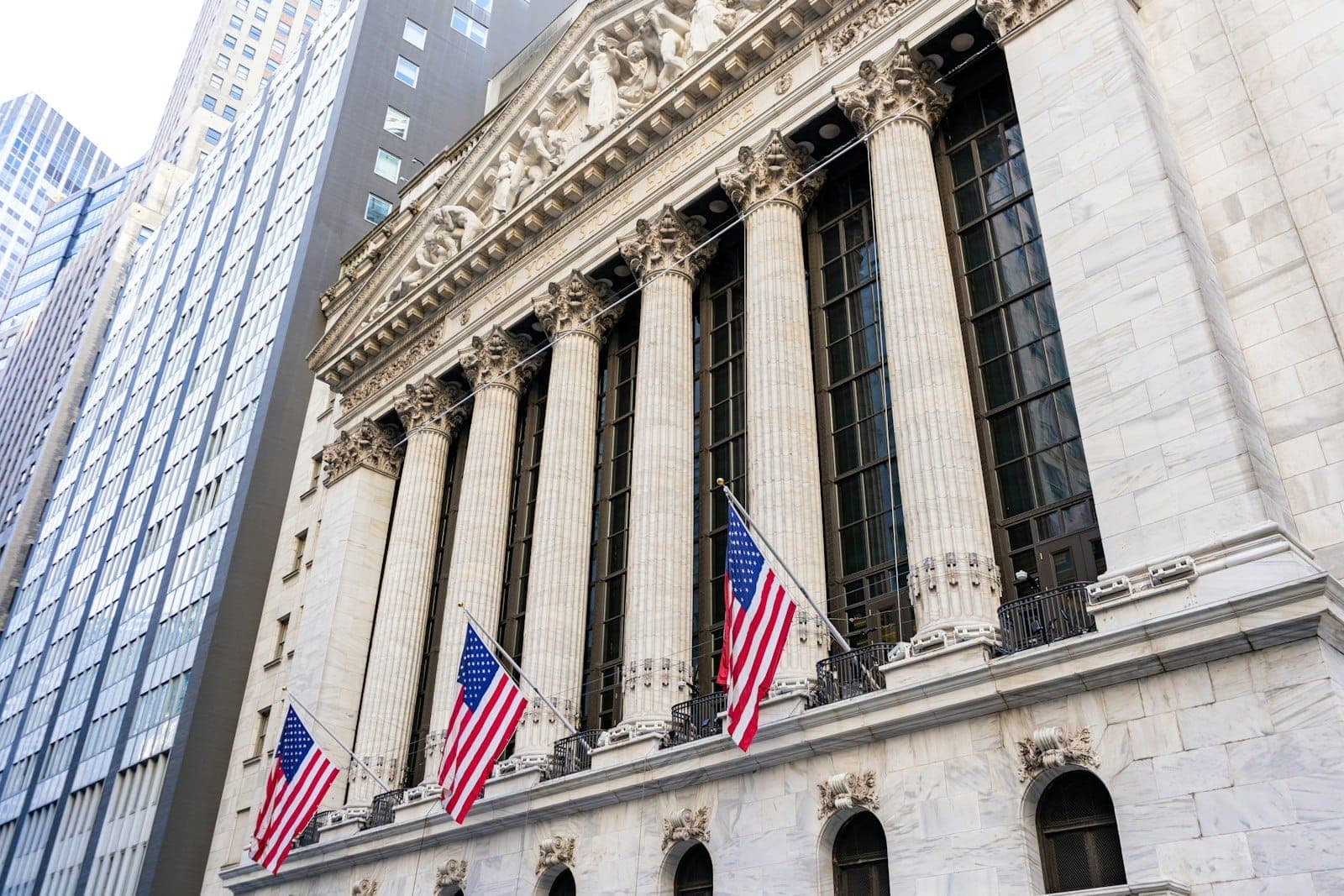 ด้านหน้าของ New York Stock Exchange บน Wall Street พร้อมเสาหินนีโอคลาสสิกขนาดใหญ่และธงชาติอเมริกันสามผืนในวันที่แดดสดใส