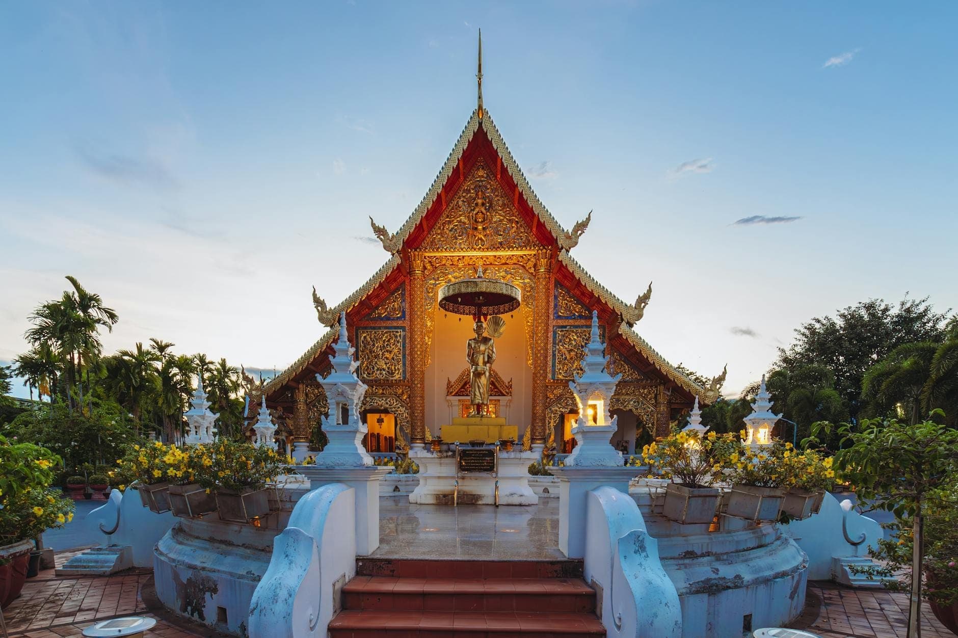 Frontansicht der Haupttempelhalle des Wat Phra Singh, reich mit Goldverzierungen geschmückt, umgeben von Grün und einem klaren blauen Abendhimmel.