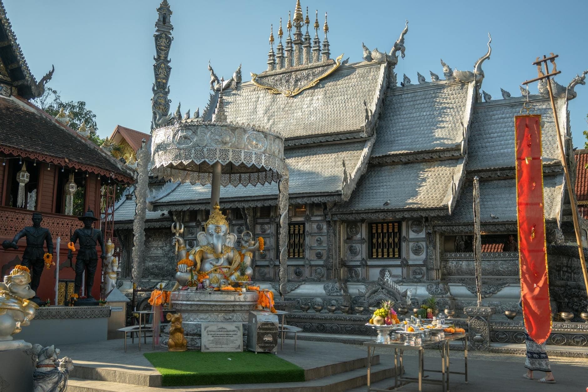 Die silberverkleidete Außenfassade von Wat Sri Suphan in Chiang Mai mit kunstvollem Metallwerk und einem Schrein davor unter klarem blauem Himmel.