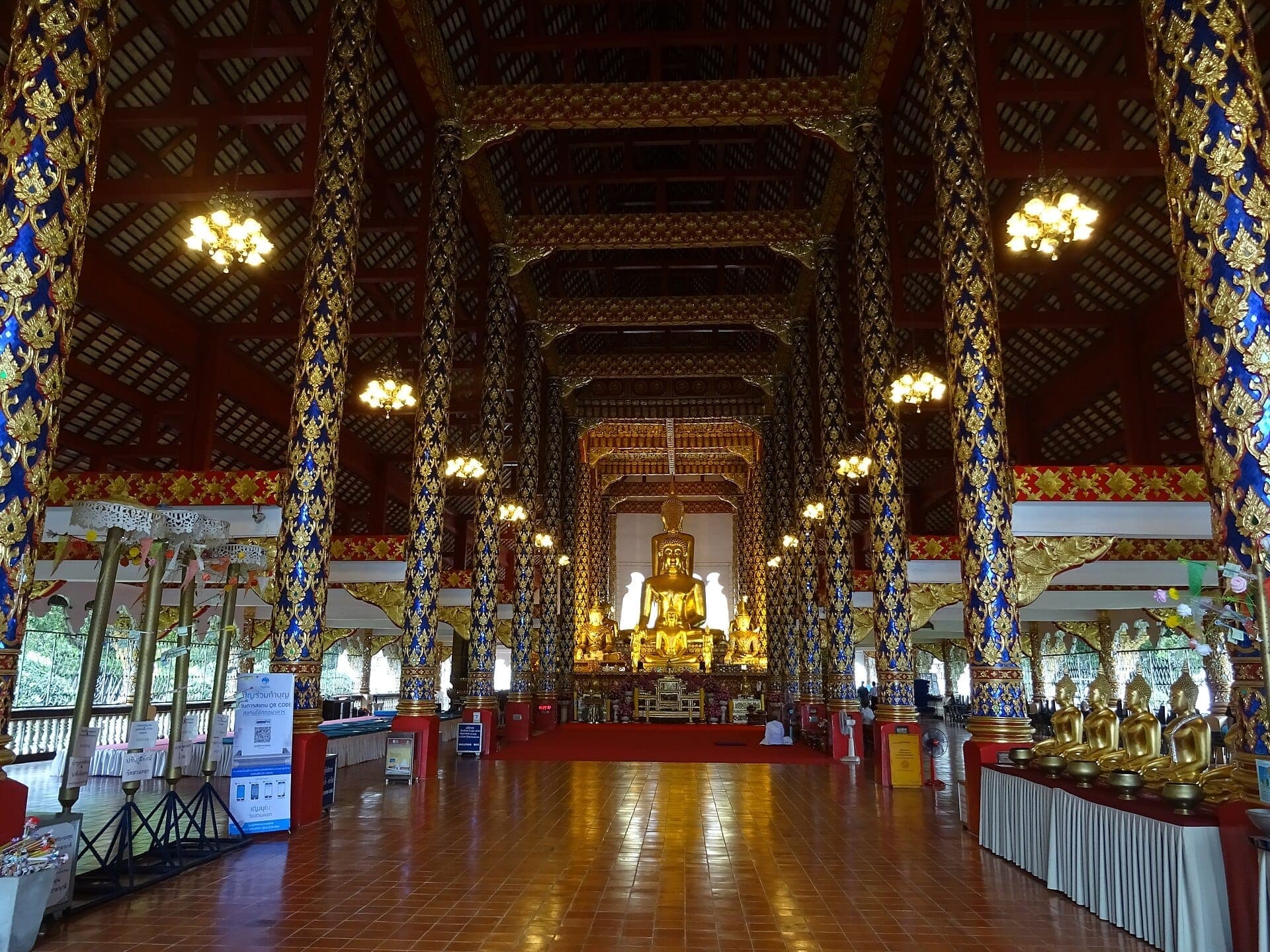 Innenansicht des Tempels Wat Suan Dok mit aufwendig dekorierten blau-goldenen Säulen, Kronleuchtern und einer großen goldenen Buddha-Statue in der Mitte.