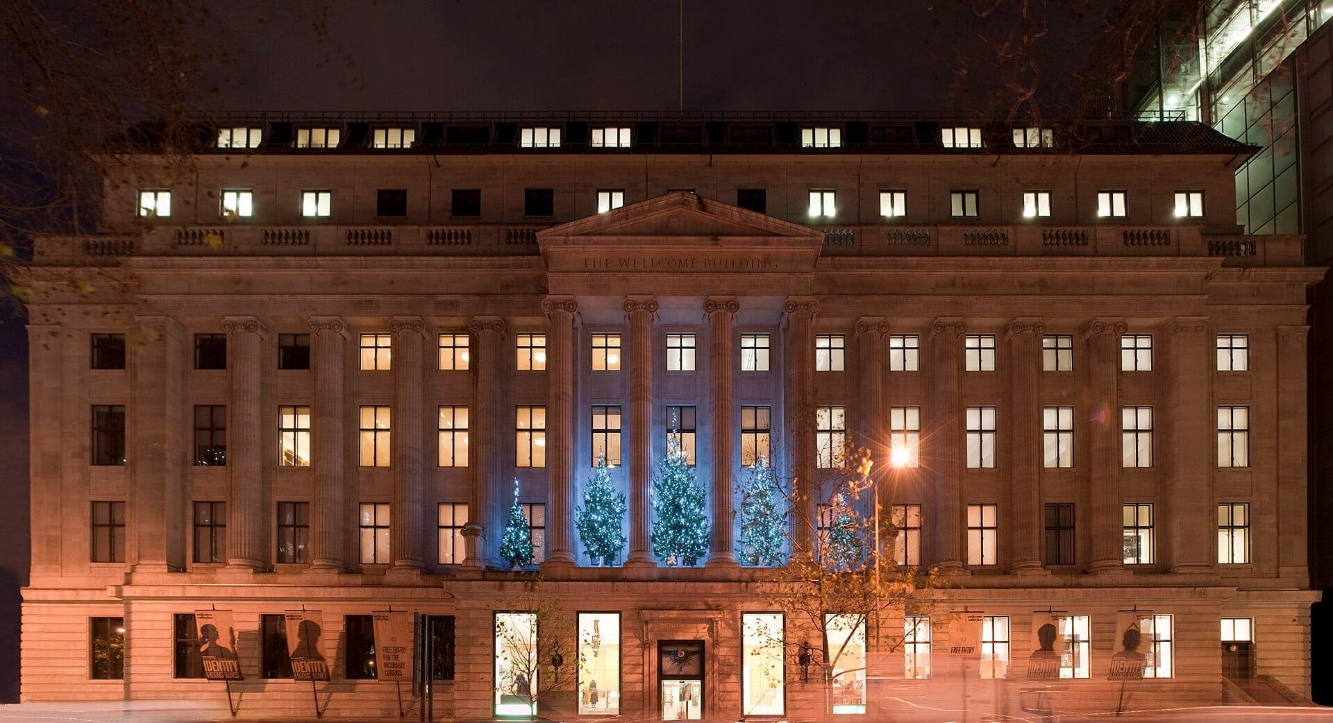 Front view of the Wellcome Collection at night, showcasing its grand neoclassical façade illuminated by warm lights and decorated with festive blue-lit trees.