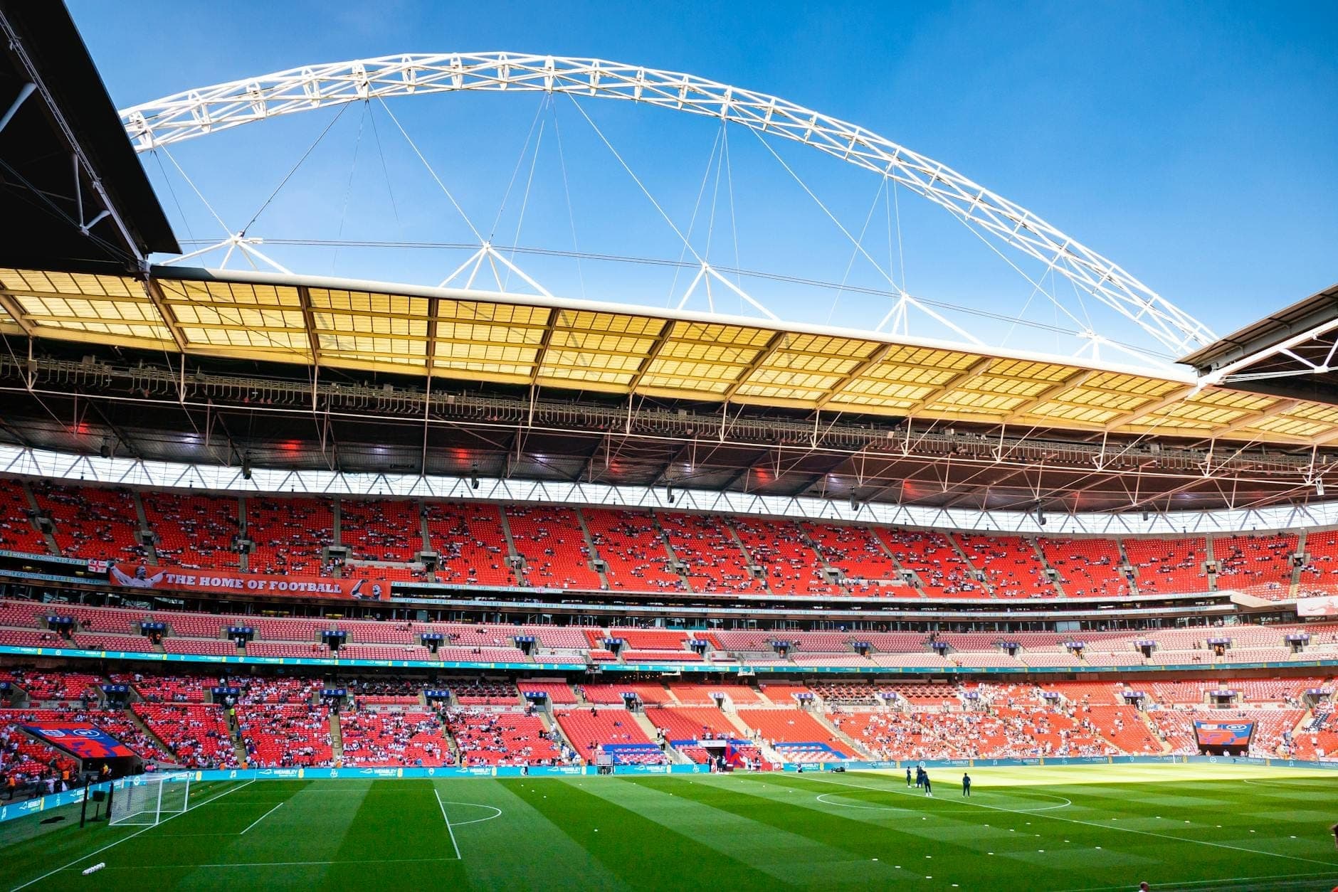 Vue grand angle de l'intérieur du stade de Wembley avec son arche blanche emblématique, sa pelouse verte et ses sièges rouges sous un ciel bleu dégagé.