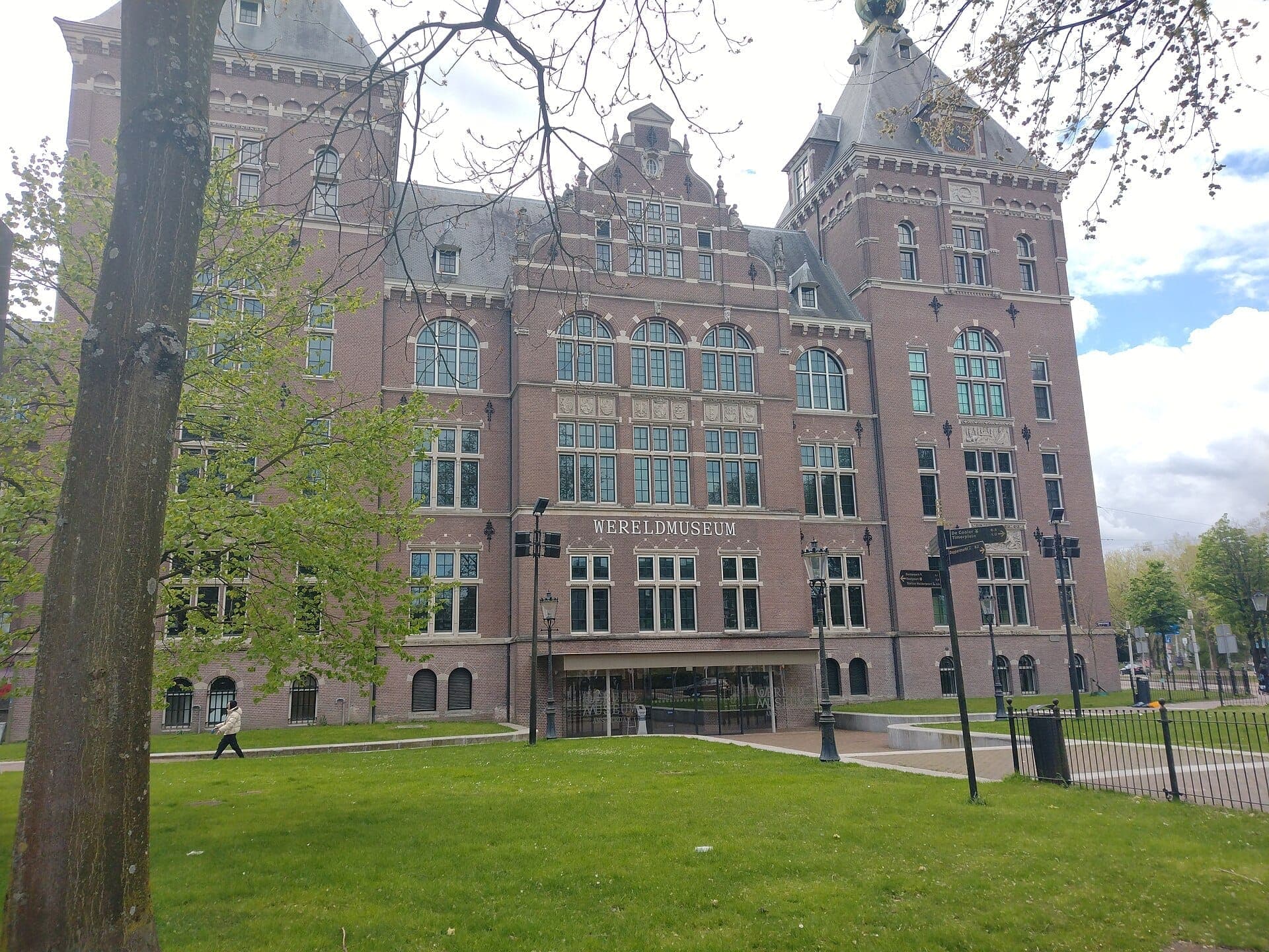 Die Fassade des Wereldmuseum Amsterdam, ein imposantes historisches Backsteingebäude mit großen Fenstern, grünen Rasenflächen und umliegenden Bäumen an einem bewölkten Tag.