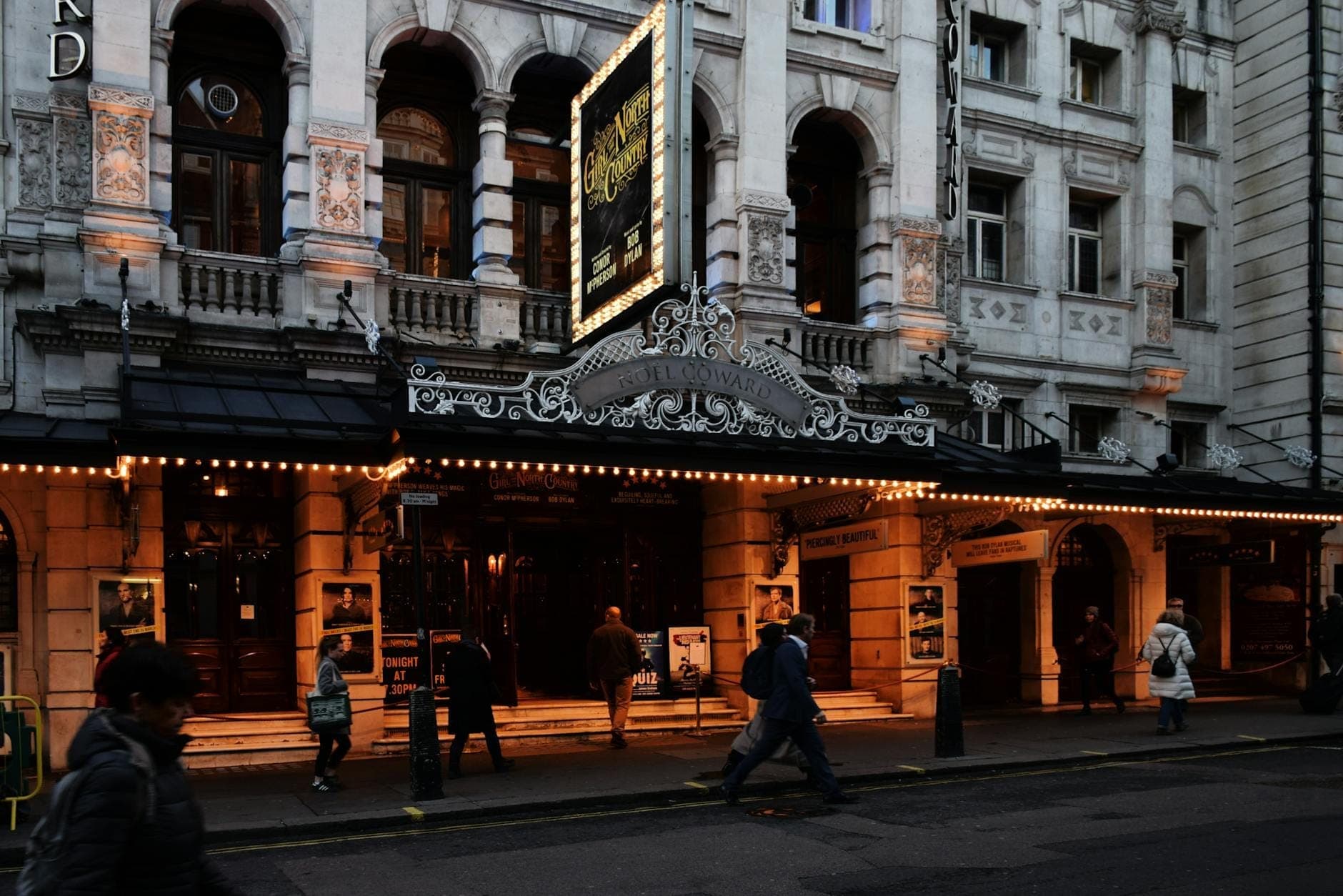 Wieczorny widok ulicy z wielkim teatrem West End w Londynie z podświetlonym szyldem i ludźmi wchodzącymi do budynku.