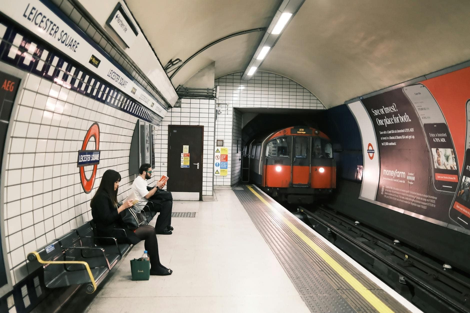 Peron londyńskiego metra na stacji Leicester Square z oczekującymi ludźmi i nadjeżdżającym pociągiem, widoczne kafelkowe ściany i czytelne oznakowanie.