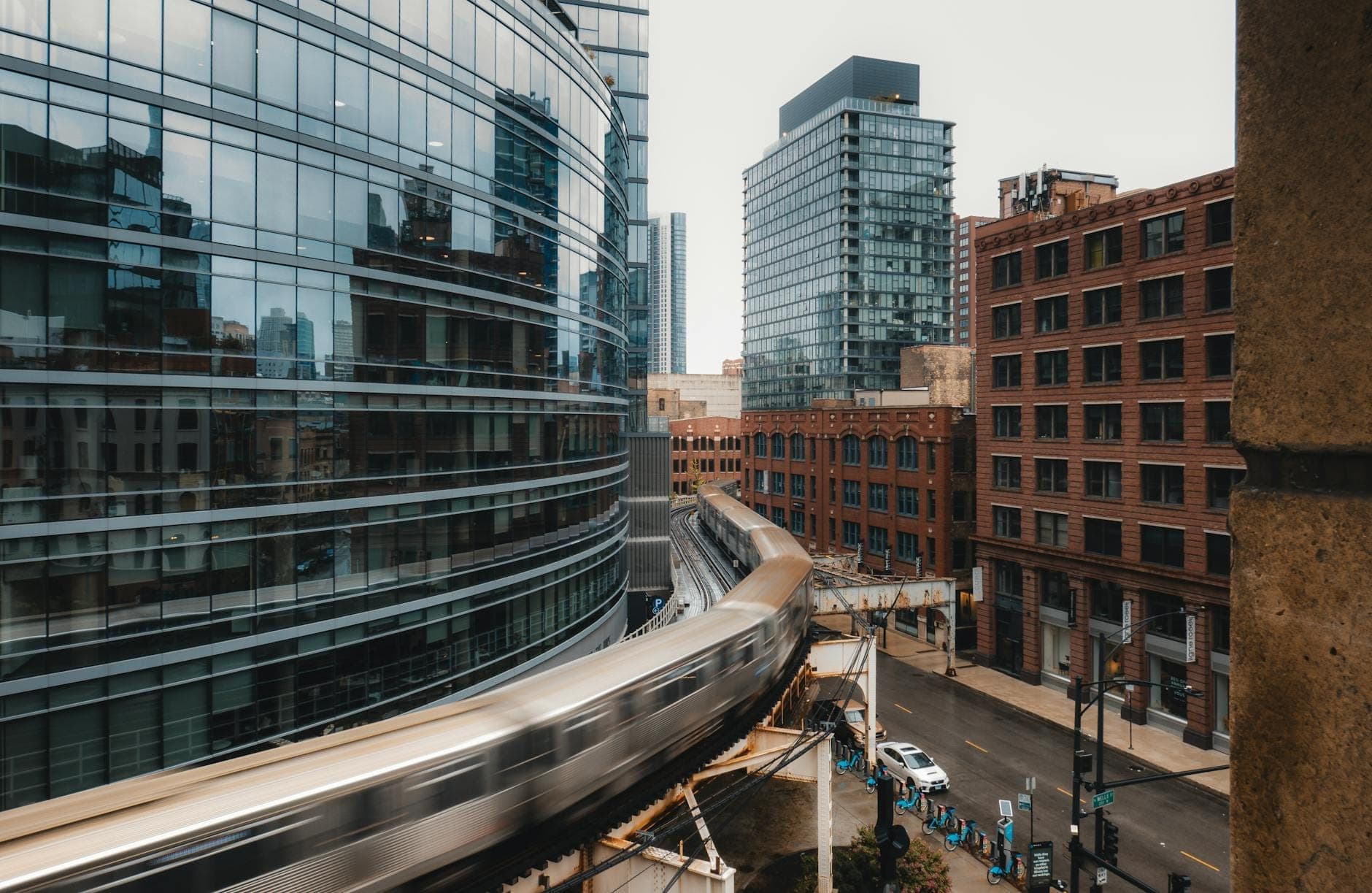 Curved elevated Chicago L train passing modern glass and historic brick buildings in West Loop with city background on an overcast day.