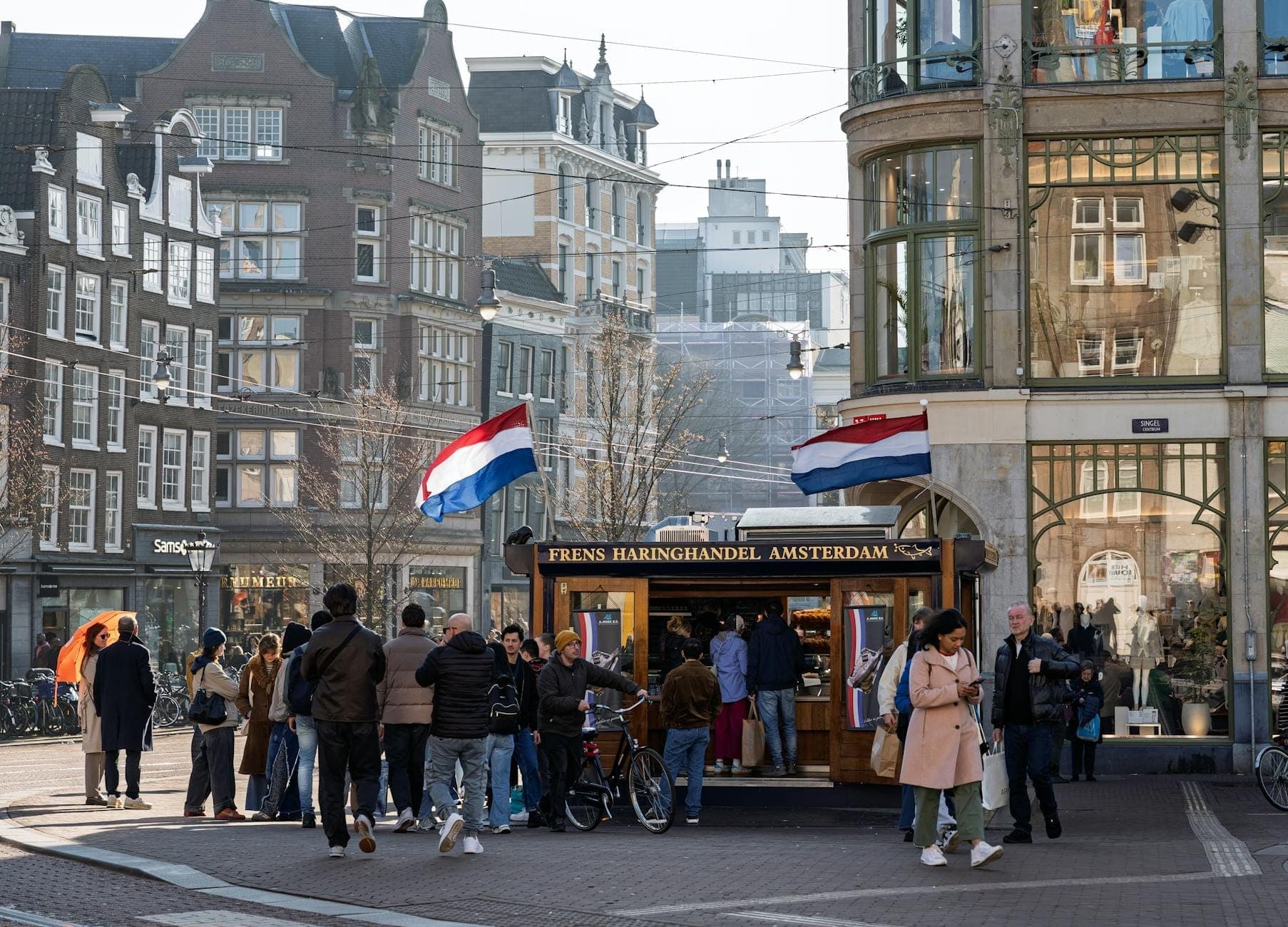 Belebte Straßenszene in Amsterdam mit Menschen, die sich an einem traditionellen Heringsstand versammeln, niederländischen Flaggen und historischen Gebäuden im Hintergrund.
