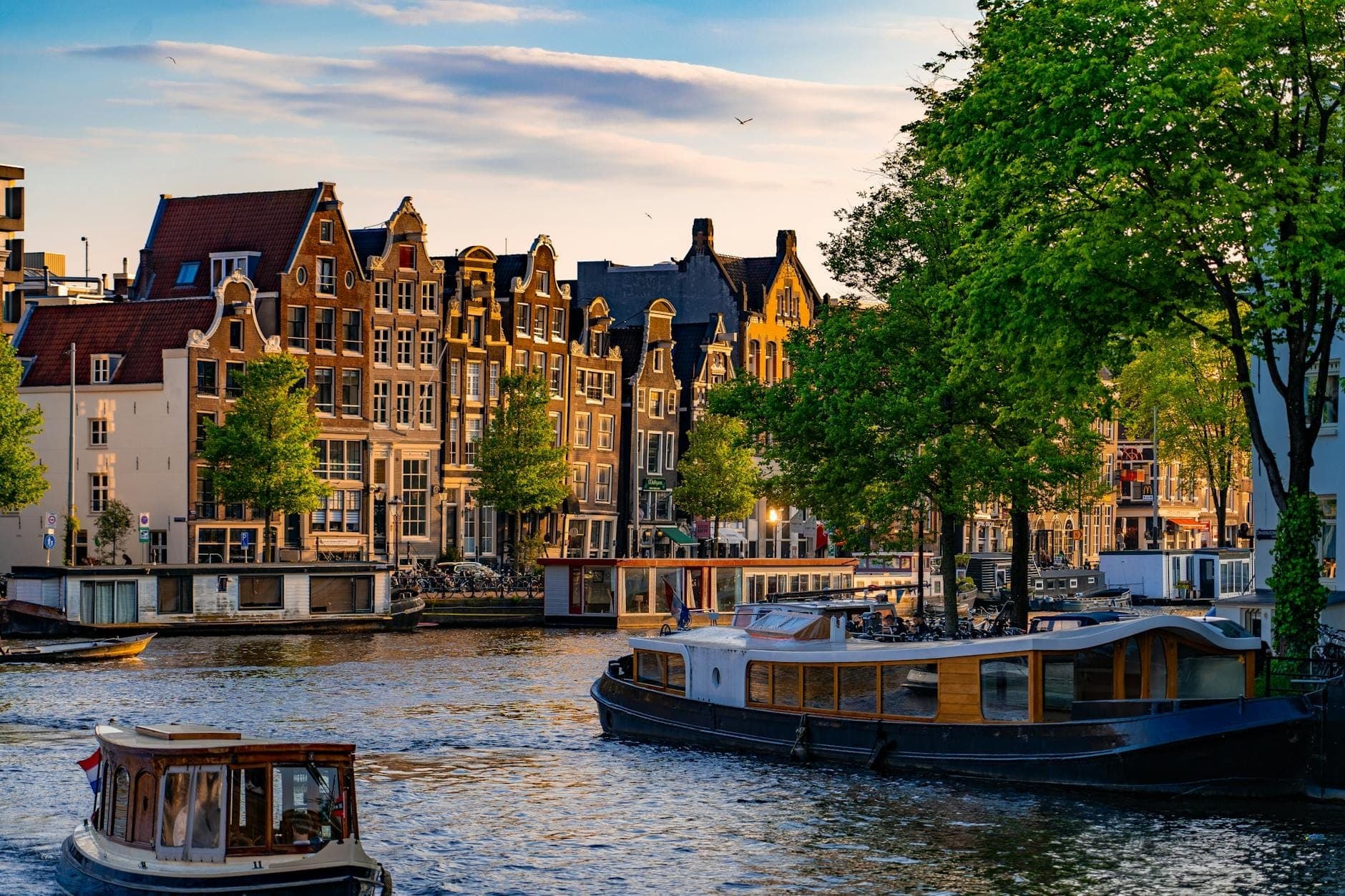 Klassischer Blick auf einen Amsterdamer Kanal mit historischen Giebelhäusern, Hausbooten und Booten unter sonnigem Himmel, der den berühmten Canal Ring der Stadt perfekt einfängt.