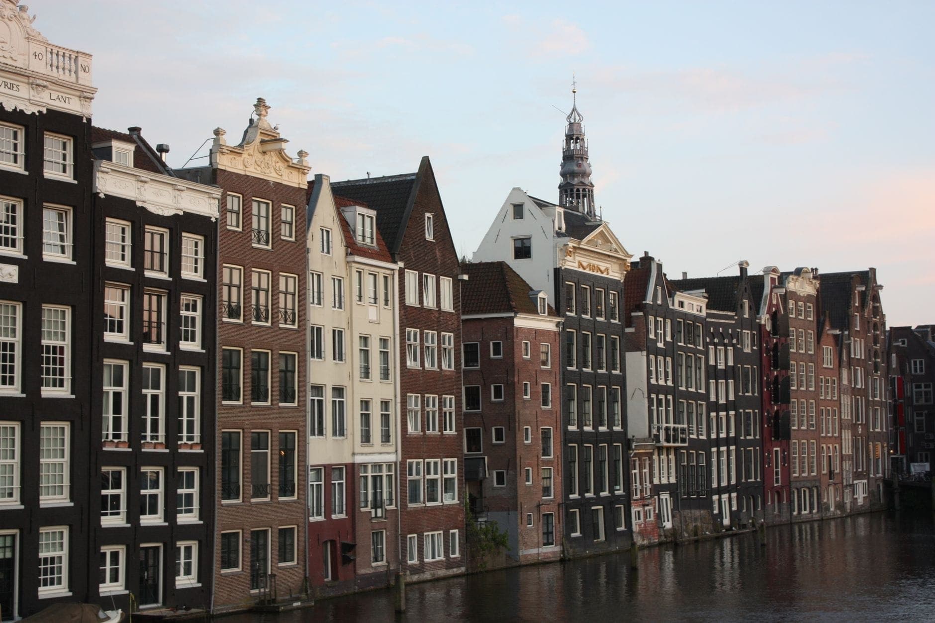 Klassische Amsterdamer Grachtenhäuser mit verzierten Giebeln und schmalen Fassaden säumen ein Ufer, mit einem Kirchturm im Hintergrund unter blauem Himmel.