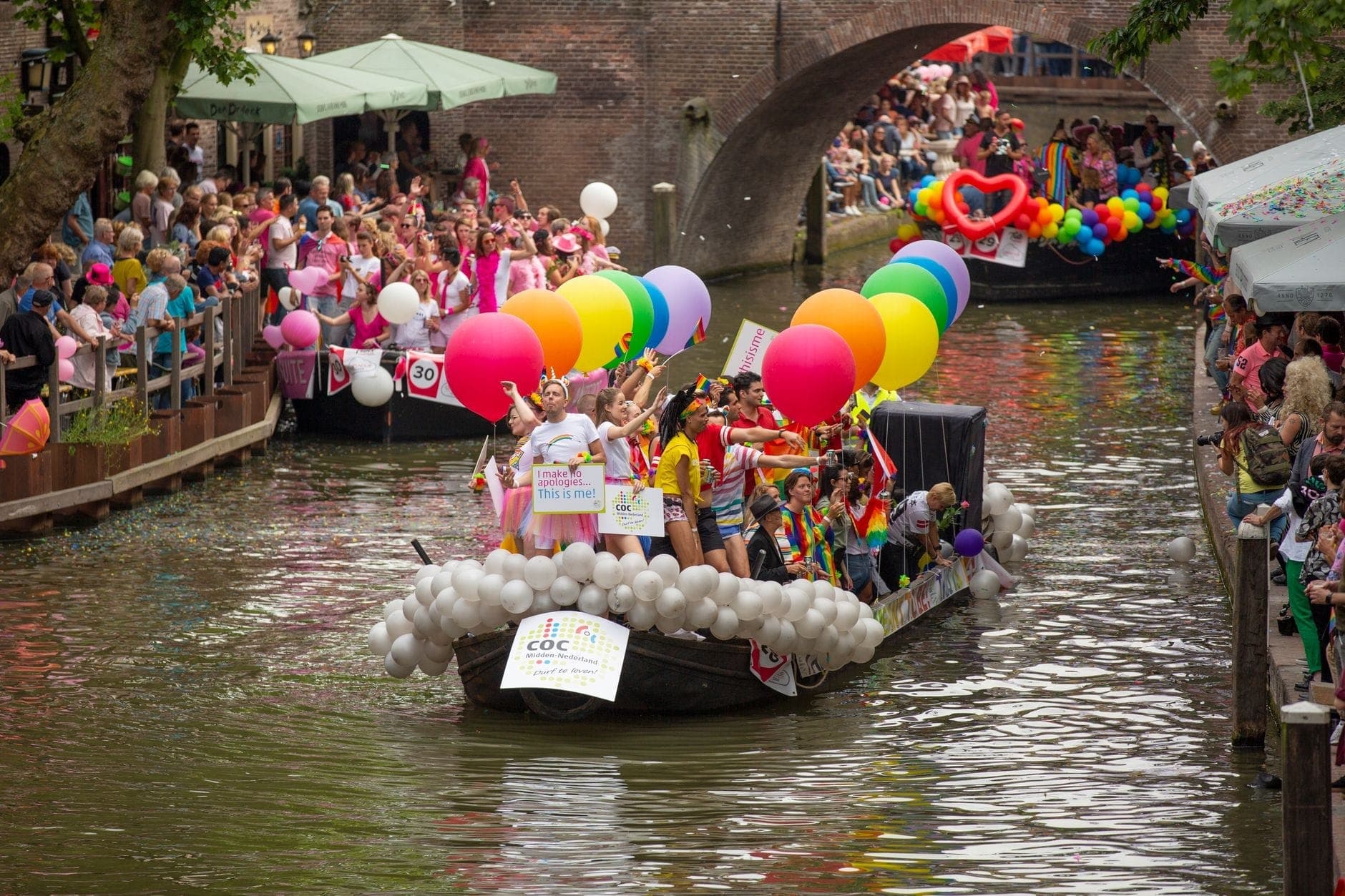 Dekoriertes Kanalboot mit Regenbogenballons und feiernden Menschen während einer Pride-Parade in Amsterdam, mit Zuschauermassen auf beiden Seiten des Kanals.
