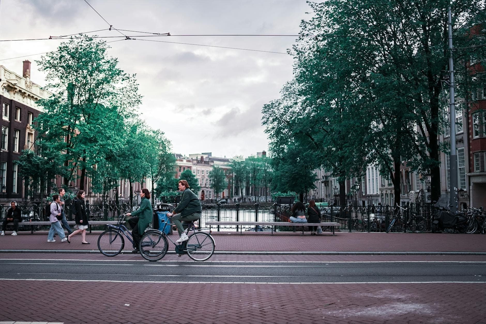 Menschen fahren Fahrrad entlang eines Kanals in Amsterdam, gesäumt von klassischen niederländischen Grachtenhäusern und Bäumen.