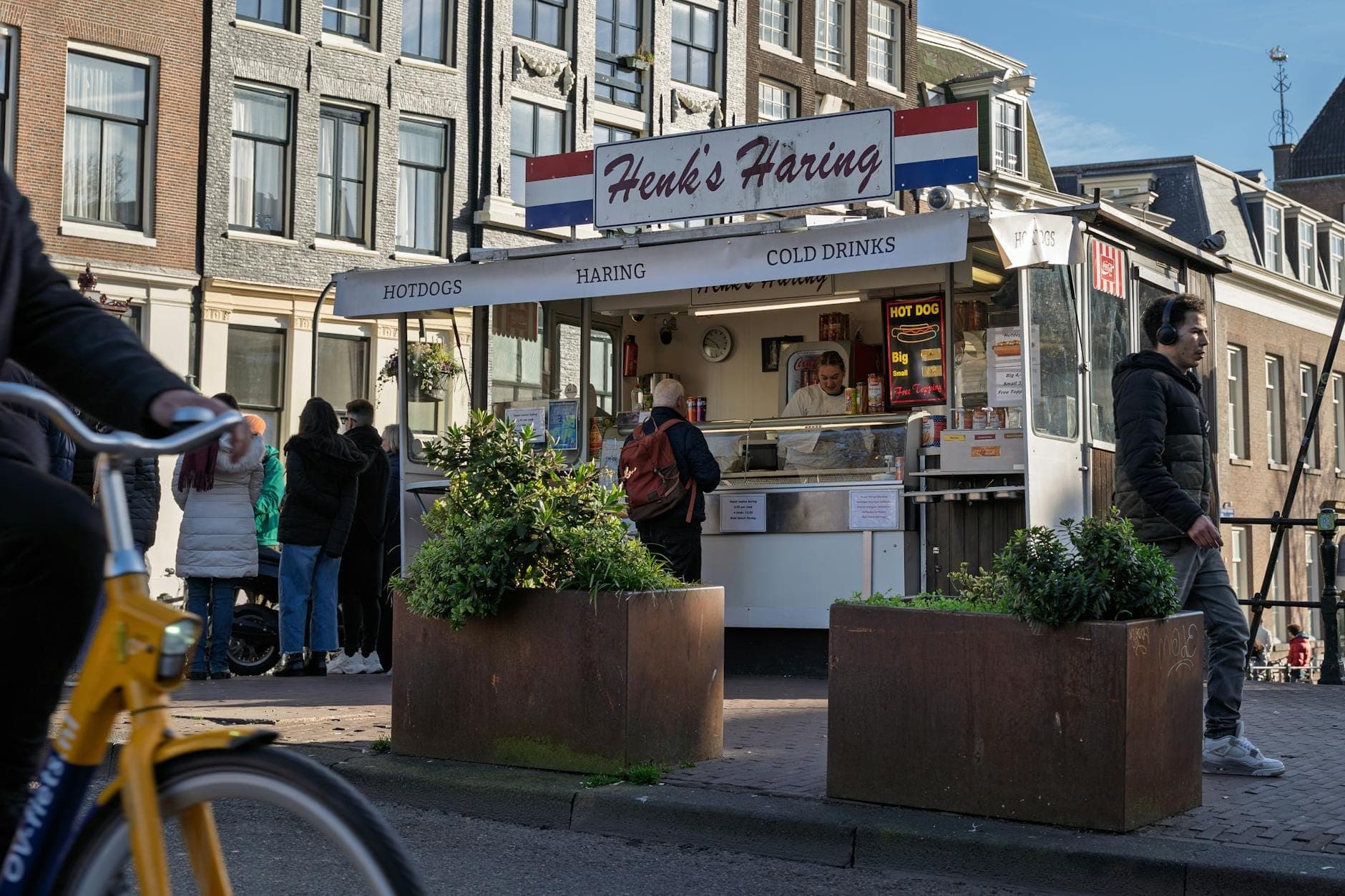 Straßenimbiss in Amsterdam mit holländischem Hering, Menschen, die Schlange stehen, typisch holländischen Gebäuden und einem vorbeifahrenden Radfahrer.