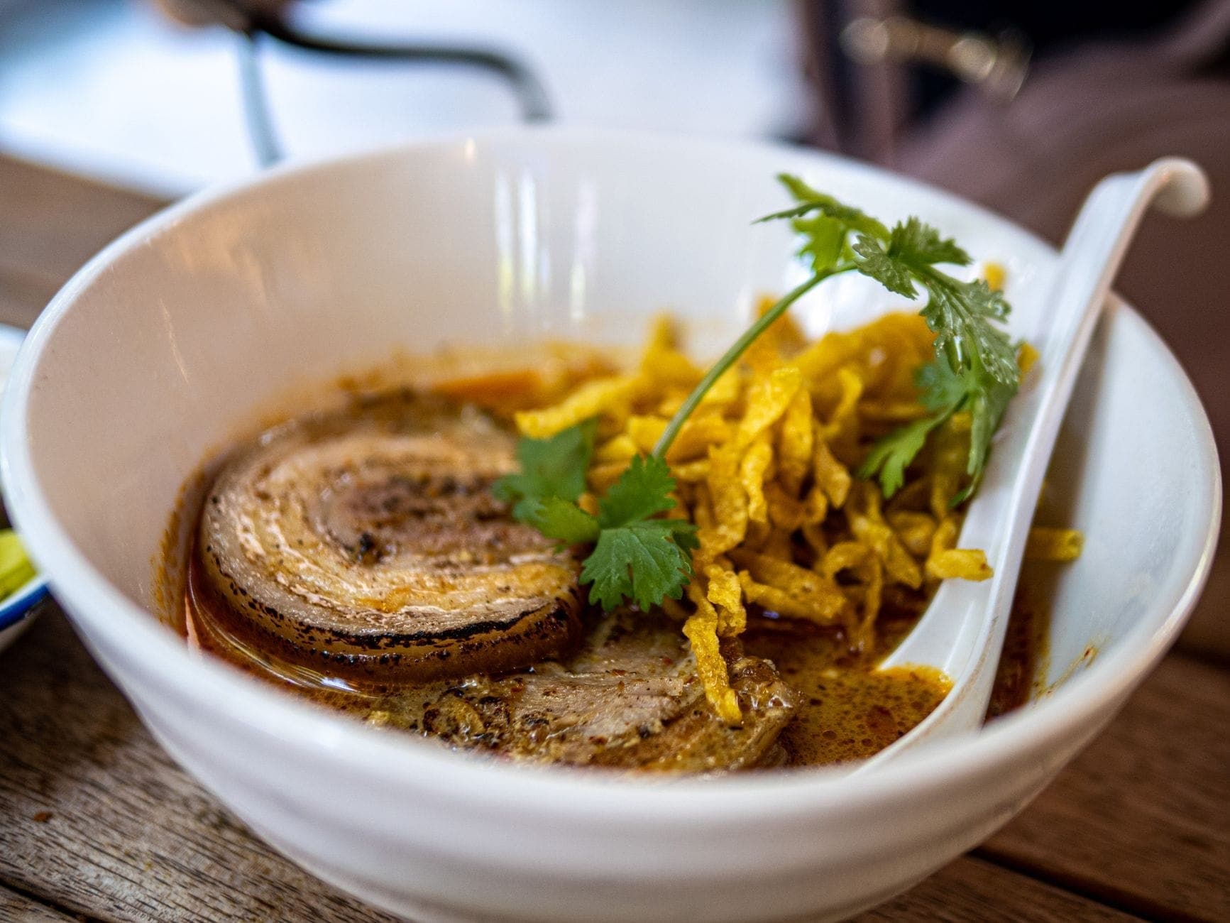 A bowl of khao soi with coconut curry broth, crispy noodles, meat, and fresh cilantro on a wooden table.