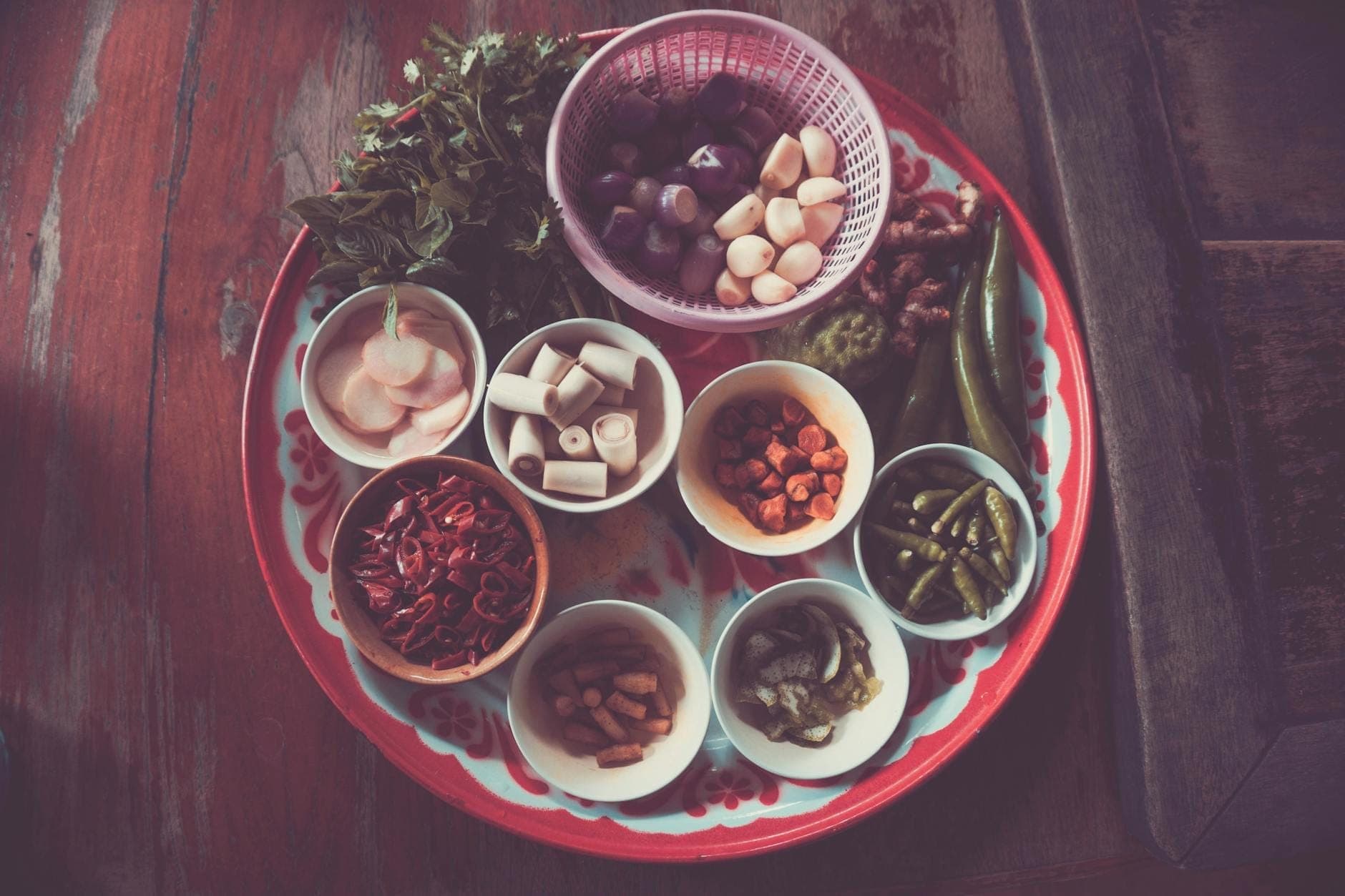 A decorative tray with small bowls of chopped herbs, spices, onions, garlic, and vegetables, ideal for a northern Thai cooking class.