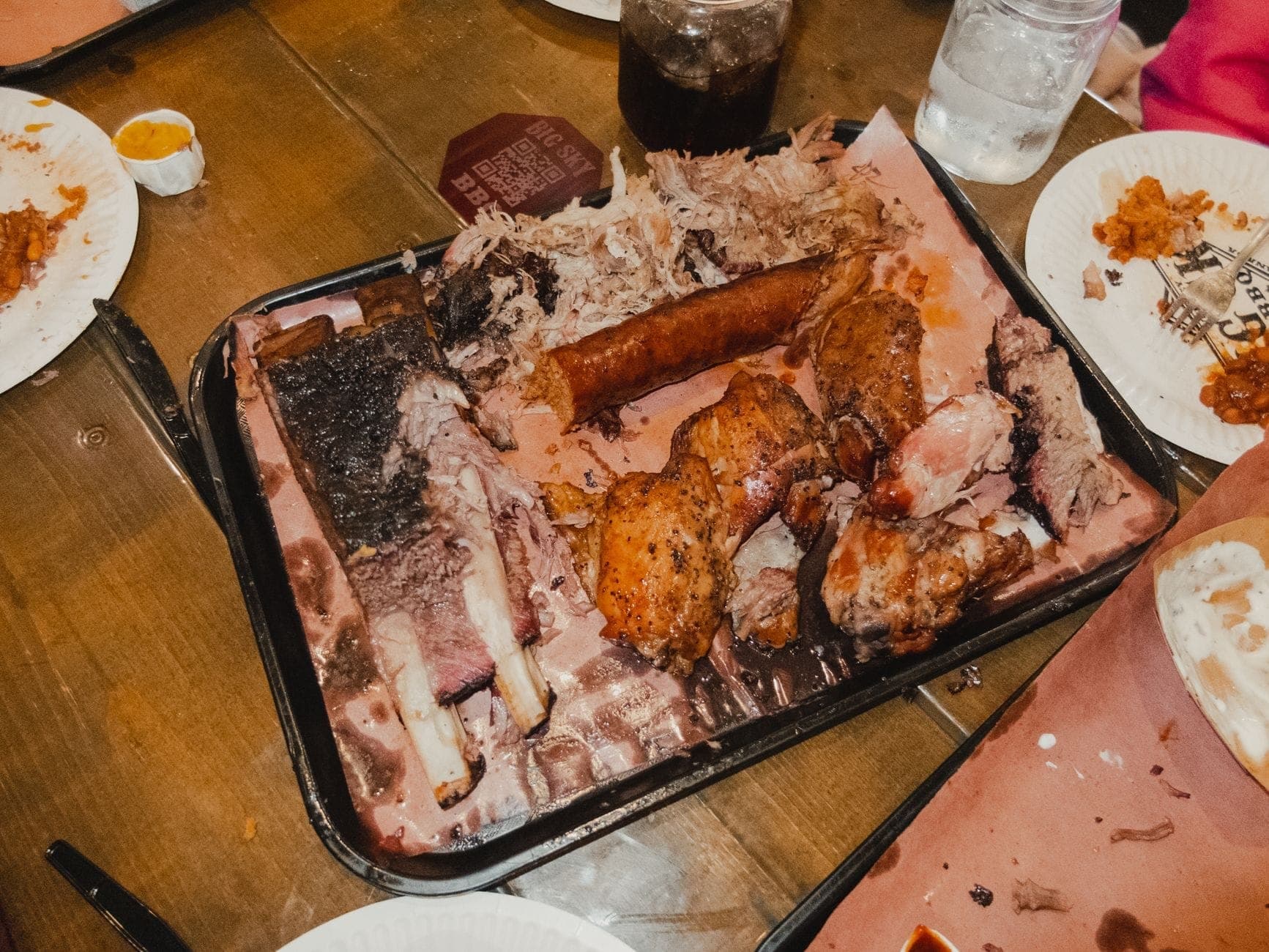 Una bandeja de carnes variadas a la barbacoa sobre papel de carnicero marrón rodeada de platos de papel y bebidas en una mesa de restaurante informal.