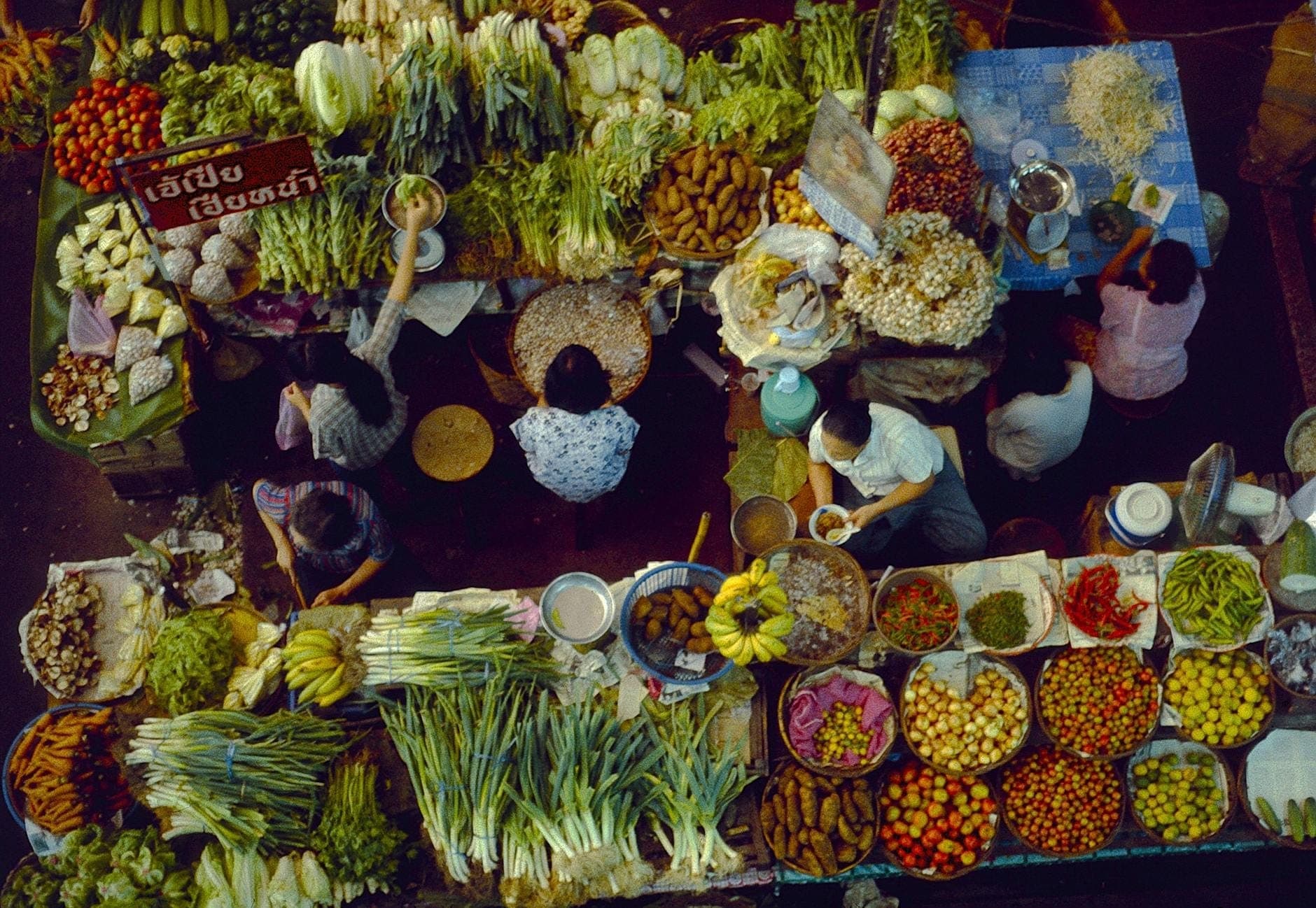 Overhead view of a busy Chiang Mai market stall with colorful fresh vegetables, fruits, and ingredients, surrounded by people preparing and selling food.