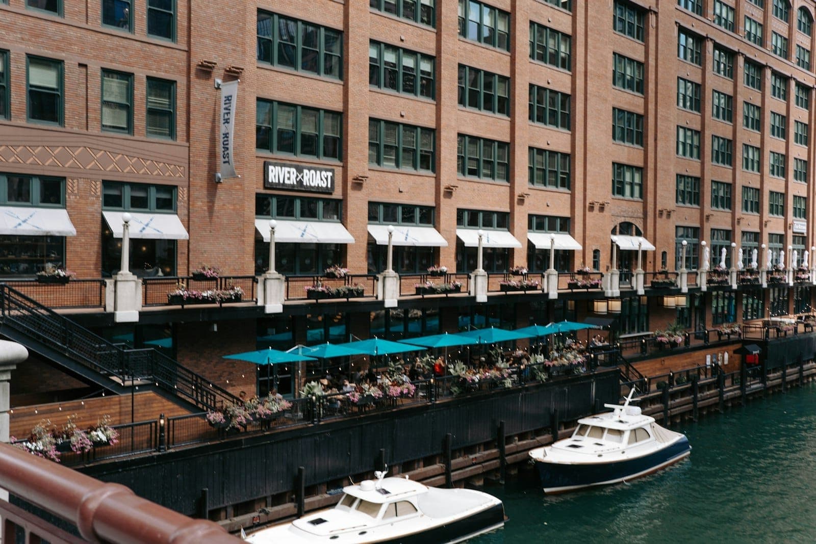 Chicago Riverwalk with outdoor restaurant seating filled with people beside the water, boats docked nearby and city buildings rising behind.