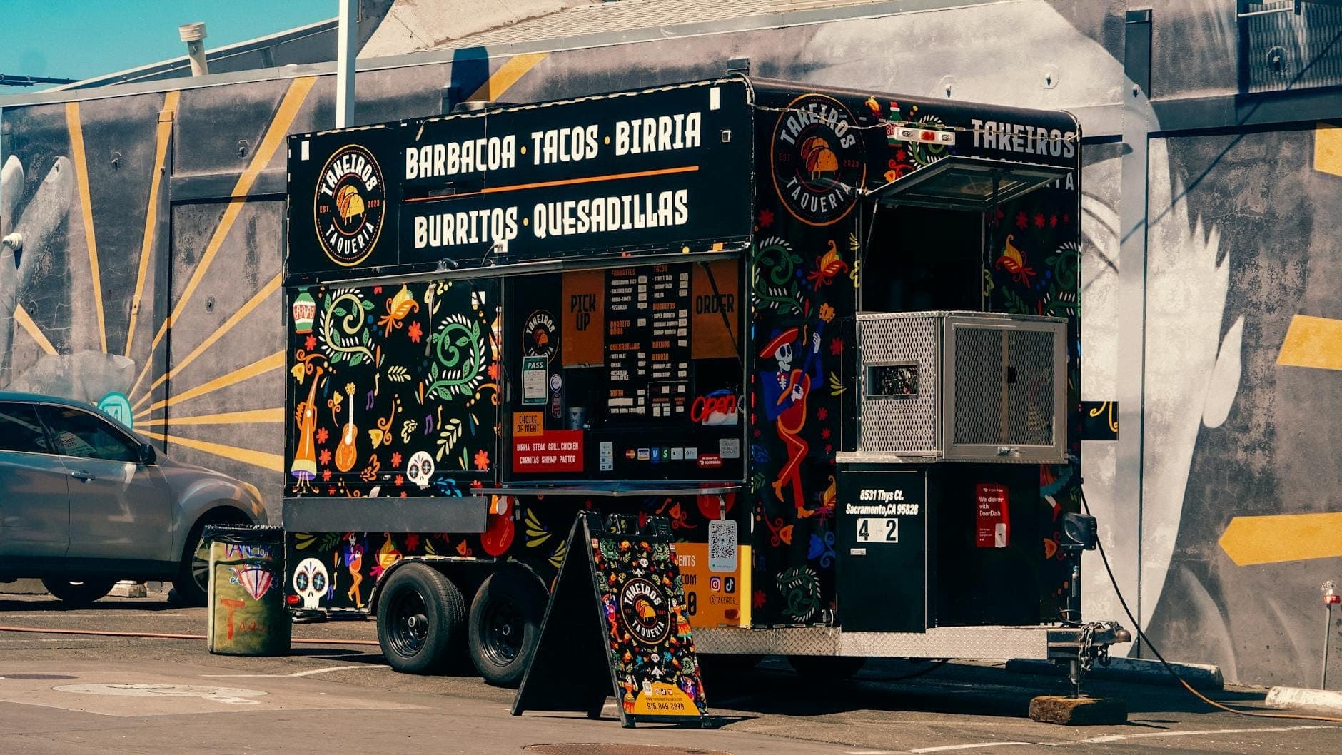 Colorful taco food truck parked in urban lot, with Mexican-inspired murals and a nearby car under bright daylight.
