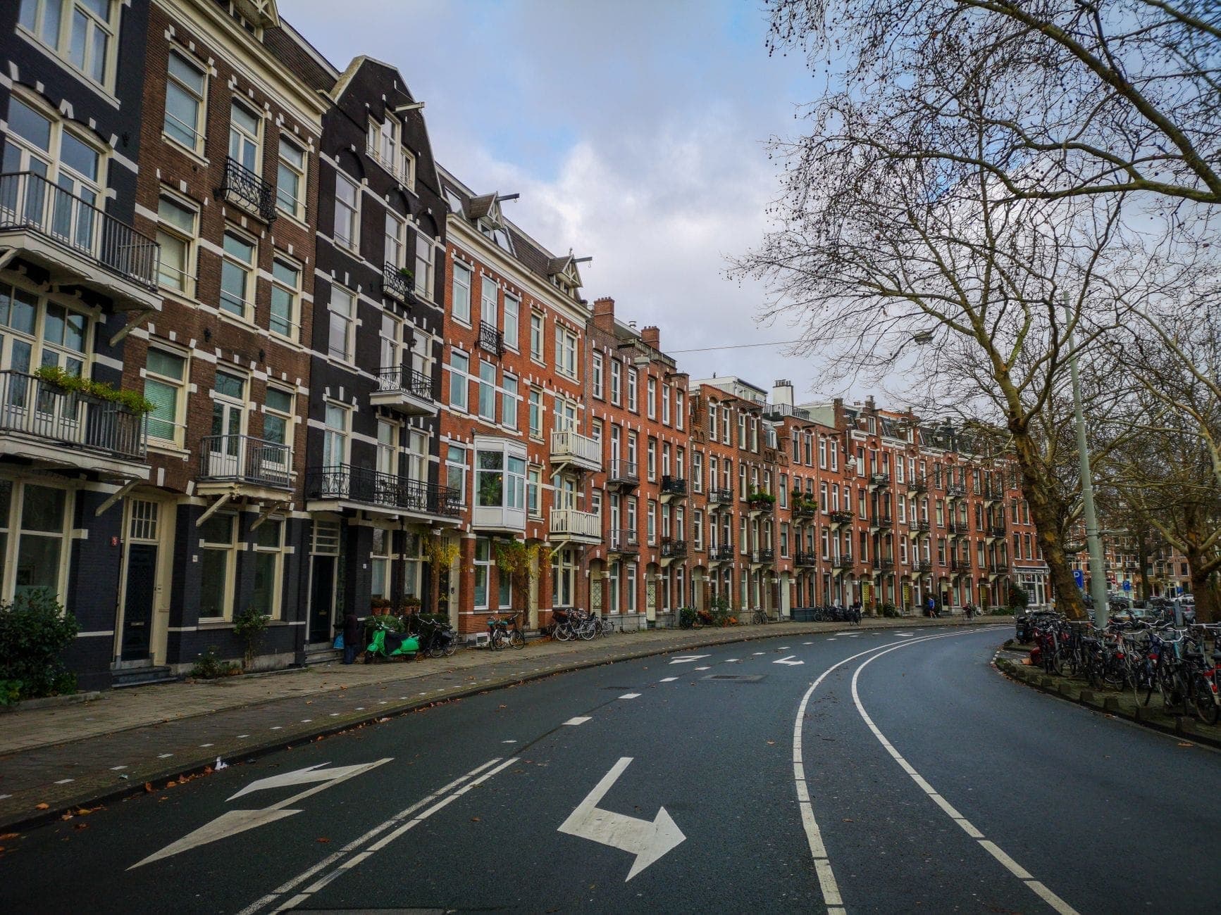 Breite Amsterdamer Straße gesäumt von traditionellen Backsteinreihenhäusern, Bäumen und auf dem Gehweg geparkten Fahrrädern an einem bewölkten Tag.