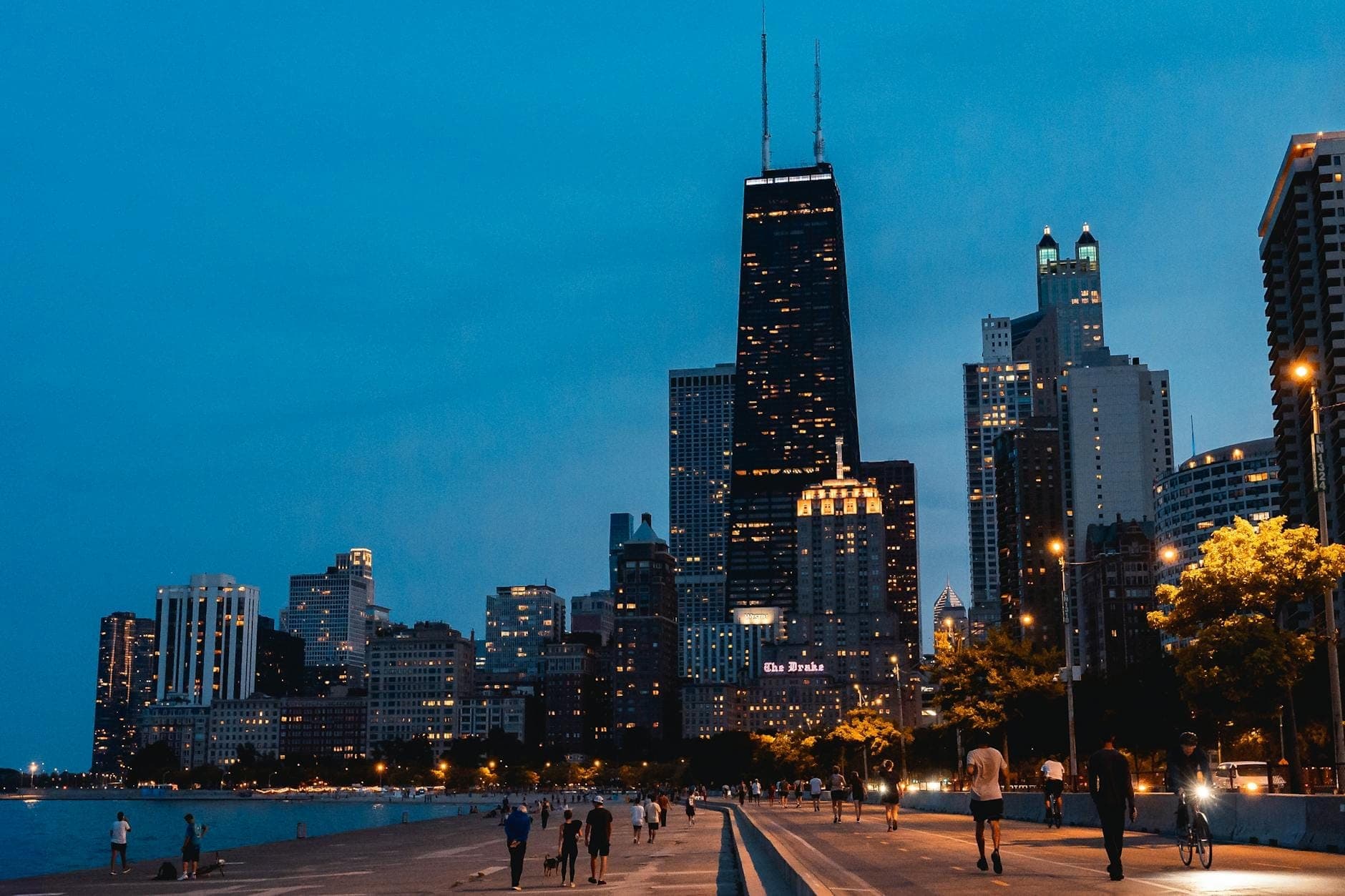 Evening view of Chicago skyline with illuminated buildings, people walking along the waterfront path, and a vibrant, welcoming urban atmosphere.