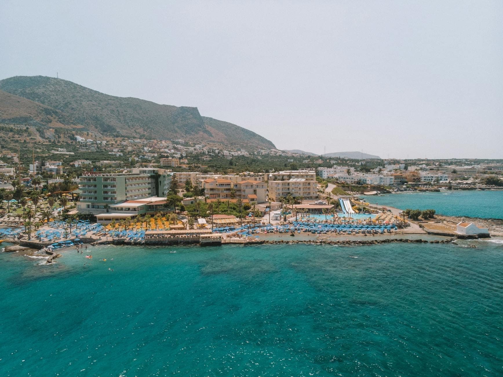 Luftaufnahme der Küste von Hersonissos mit großen Hotels, Liegestühlen, Pools und einer lebhaften Uferpromenade, typisch für Kretas Ferienstreifen.