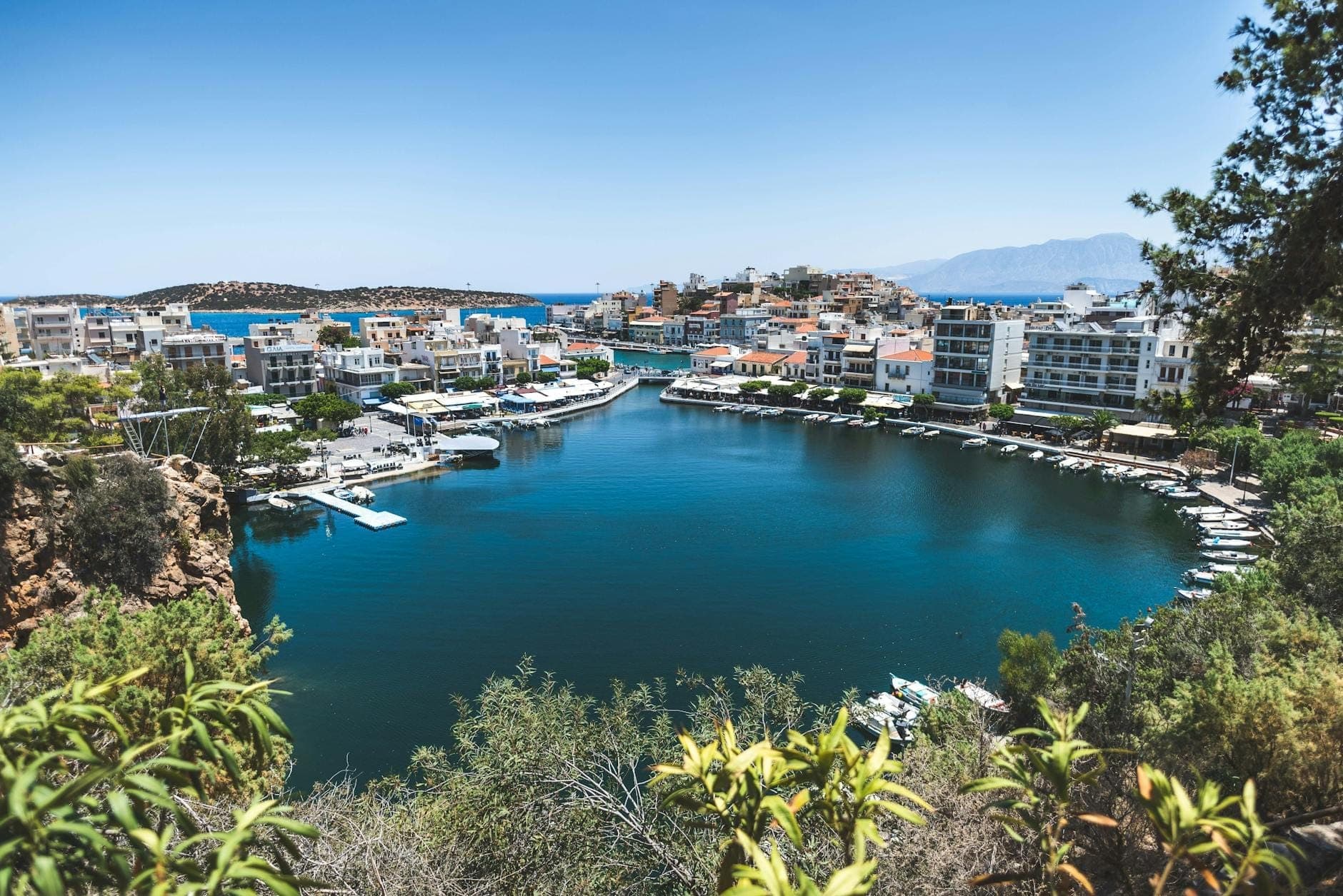 Malerische Küstenstadt auf Kreta mit weiß-pastellfarbenen Häusern rund um eine blaue Lagune, kleinen Booten und Bergen unter klarem Himmel.