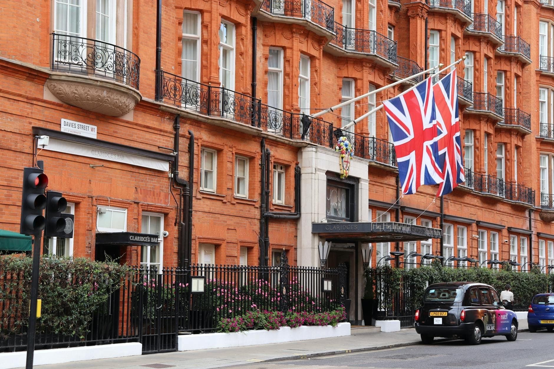 Elegante Backsteinfassade des Hotels Claridge's in Mayfair, London, mit Union-Jack-Flaggen und einem schwarzen Londoner Taxi davor.