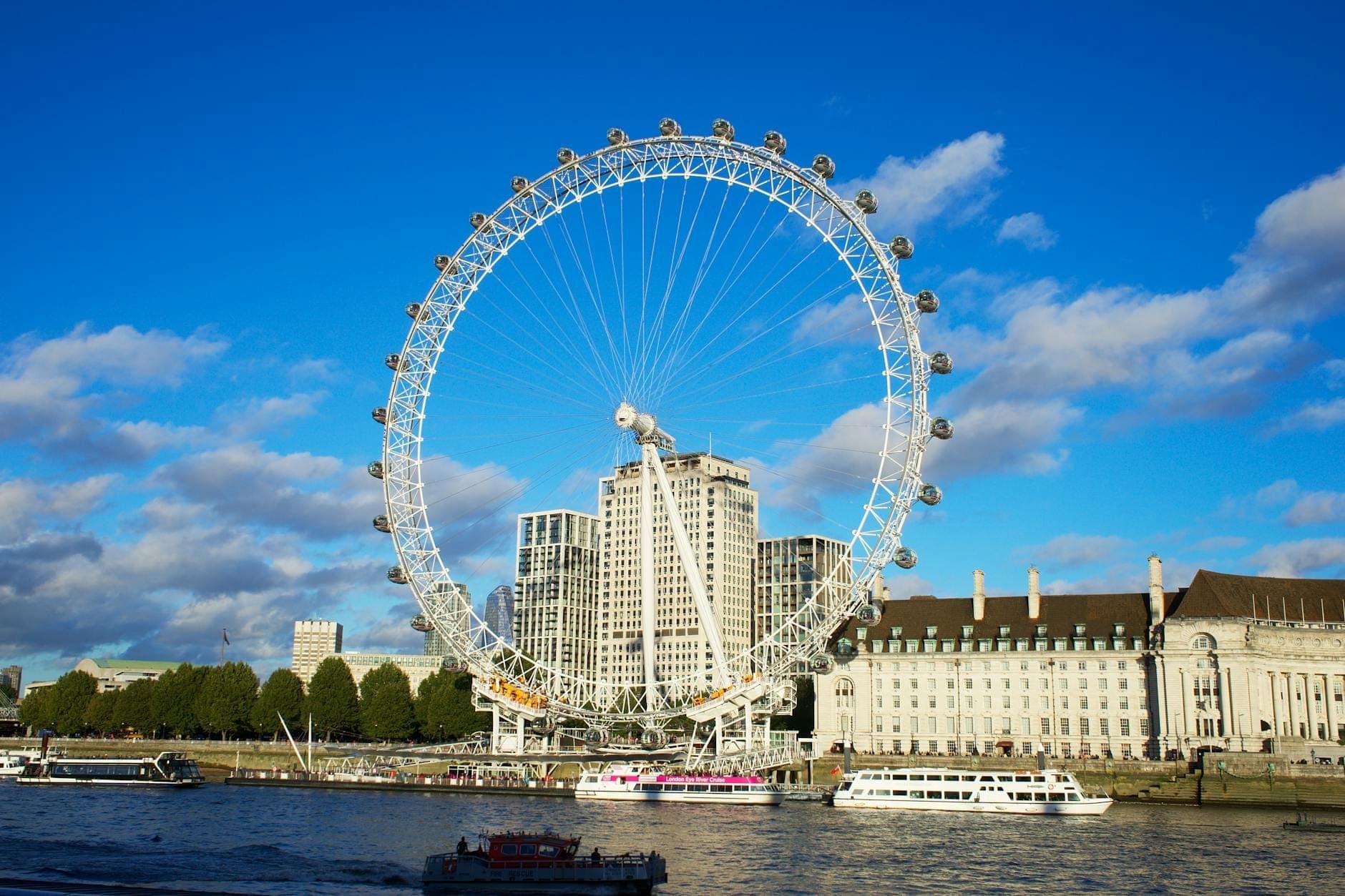 Das London Eye am Südufer neben der Themse, unter einem strahlend blauen Himmel mit Booten auf dem Fluss.