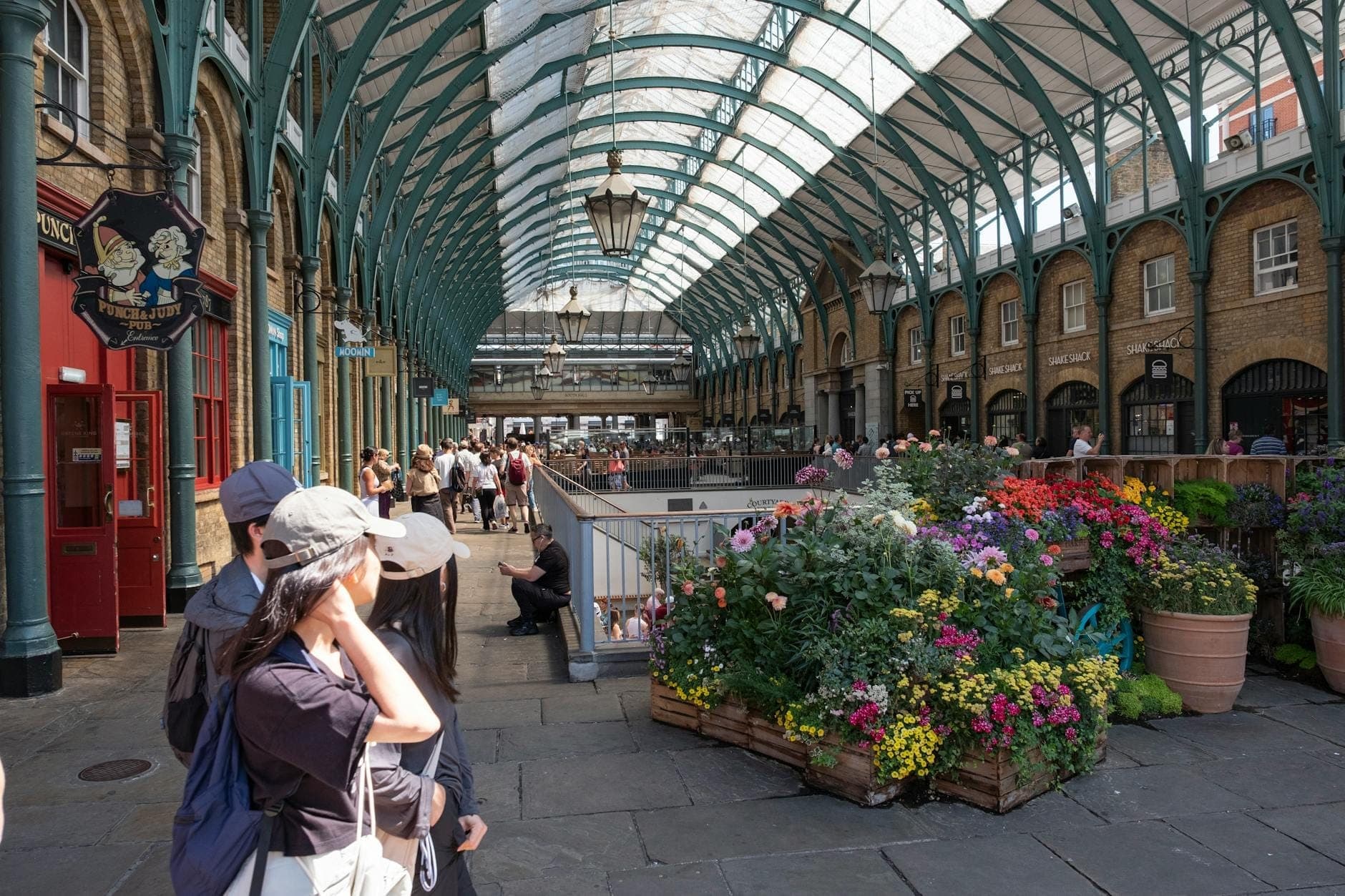 Weitwinkelperspektive des Covent Garden Market mit Glasdach, Blumendekorationen und Menschen, die die Markthalle im Londoner West End erkunden.