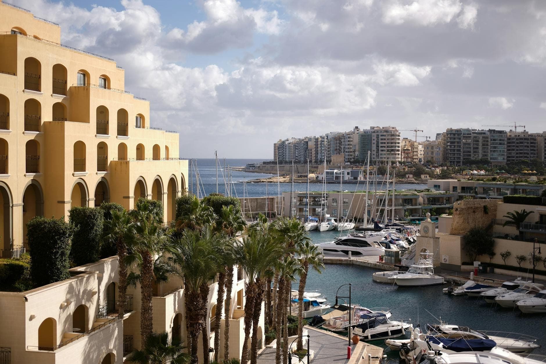Elegante Gebäude am Wasser mit Palmen und einer Marina voller Yachten in Malta, im Hintergrund Stadtsilhouette und Meer.