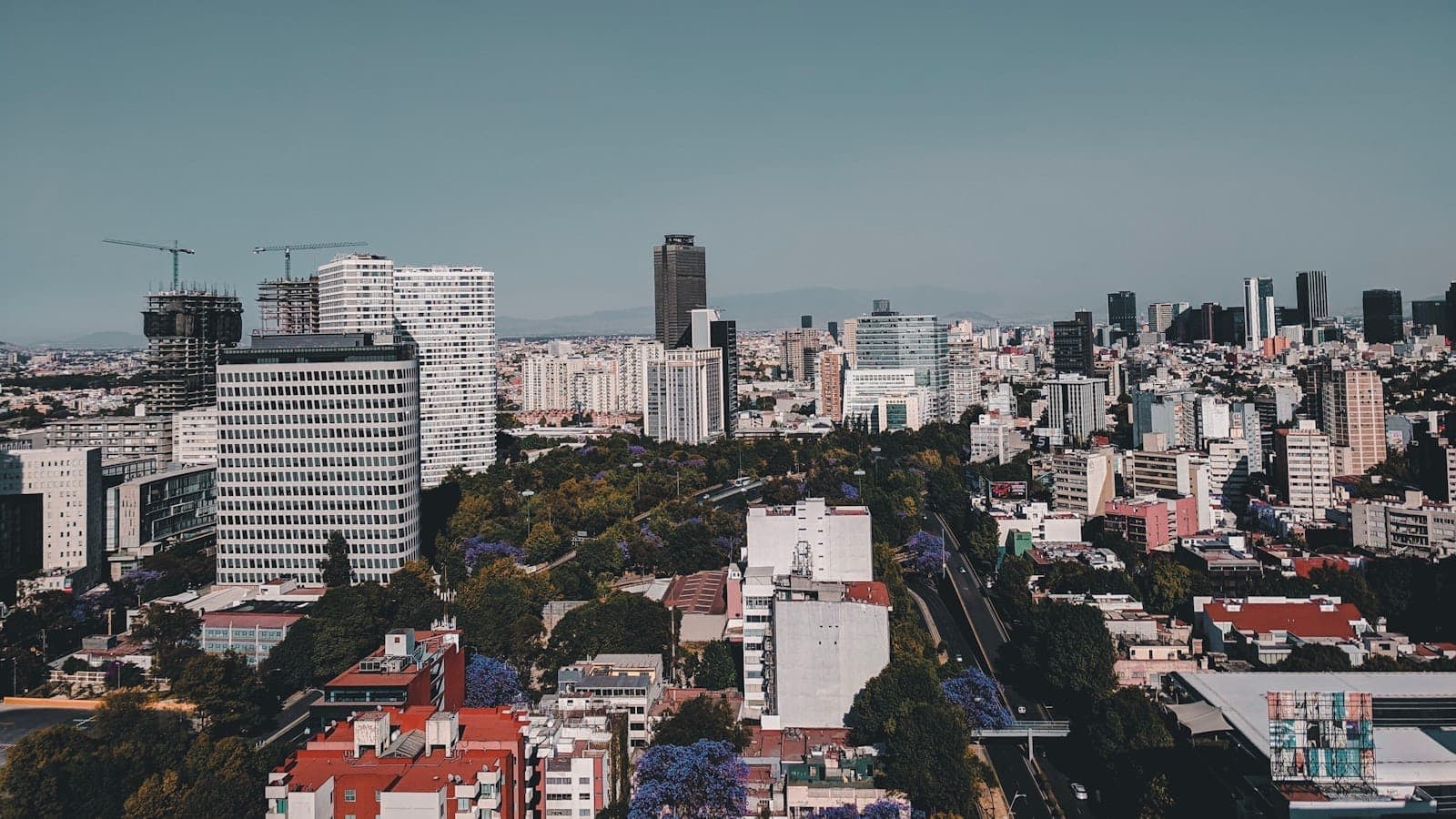 Aerial view of Mexico City with modern high-rise buildings, residential areas, and tree-lined streets under a clear sky.