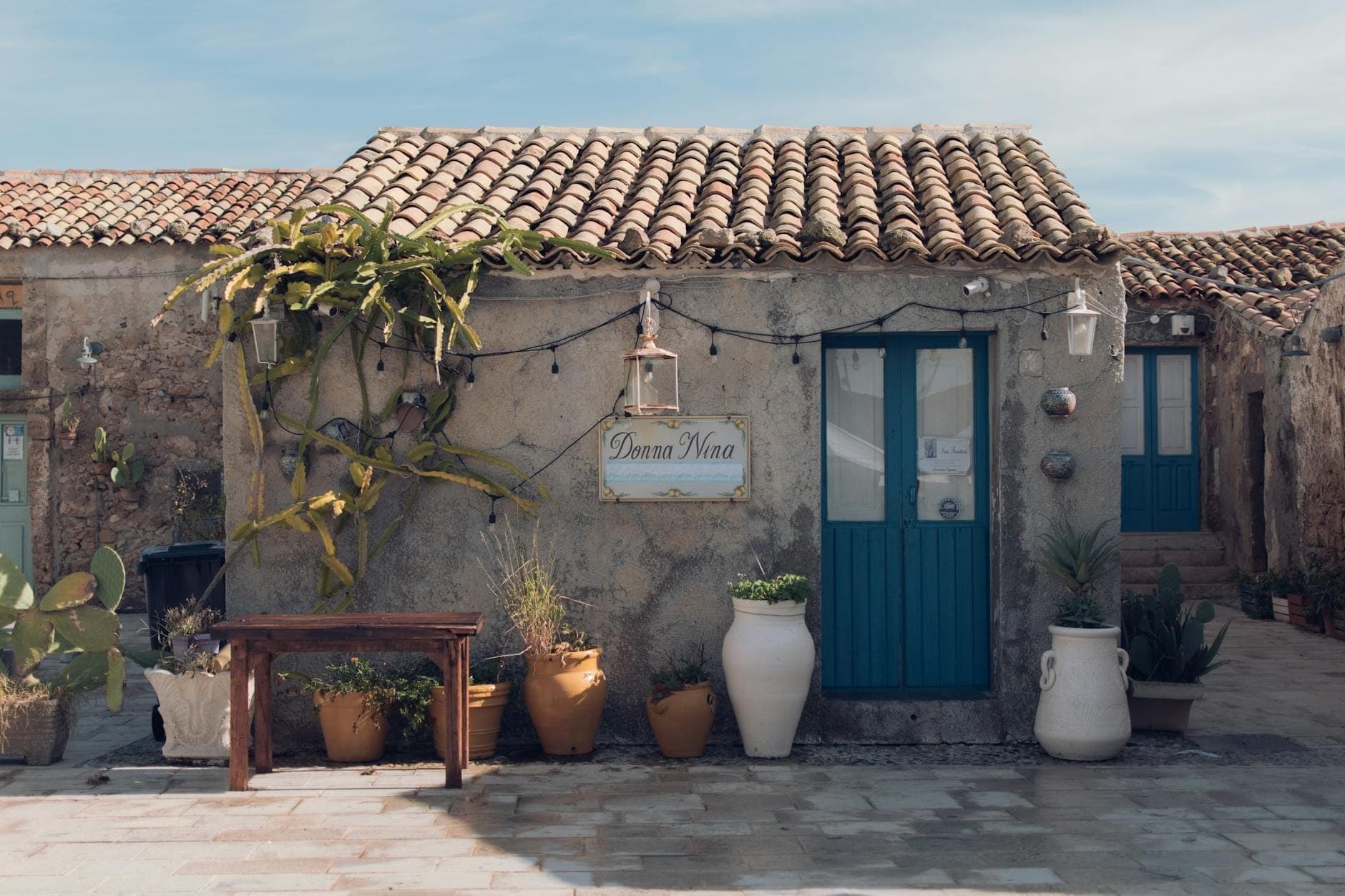 Casa de huéspedes rústica en Sardegna con paredes de piedra, puertas azules, plantas en macetas y un letrero, que representa el estilo de alojamiento local.