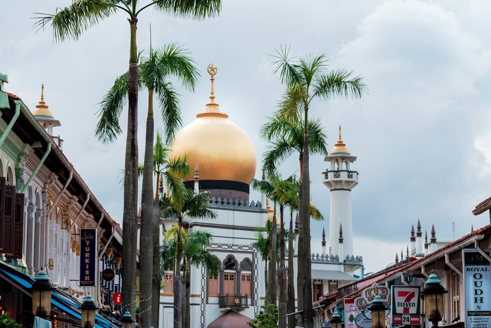 วิวของ Sultan Mosque พร้อมต้นปาล์มและอาคารพาณิชย์ใน Kampong Glam สิงคโปร์ ใต้ท้องฟ้าที่มีเมฆมาก