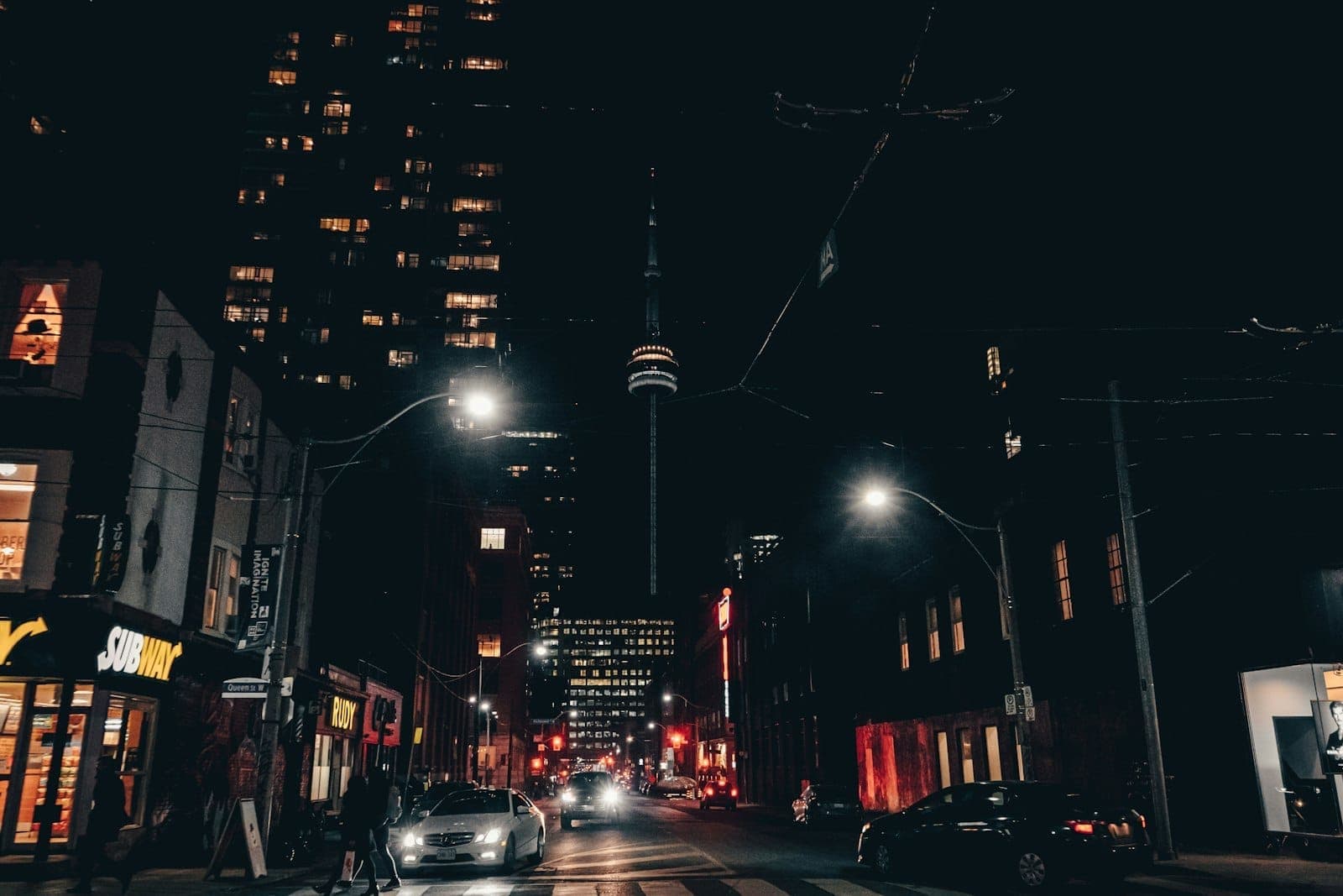 Nighttime street view in downtown Toronto with the CN Tower visible, surrounded by lit-up bars, restaurants, and city buildings.