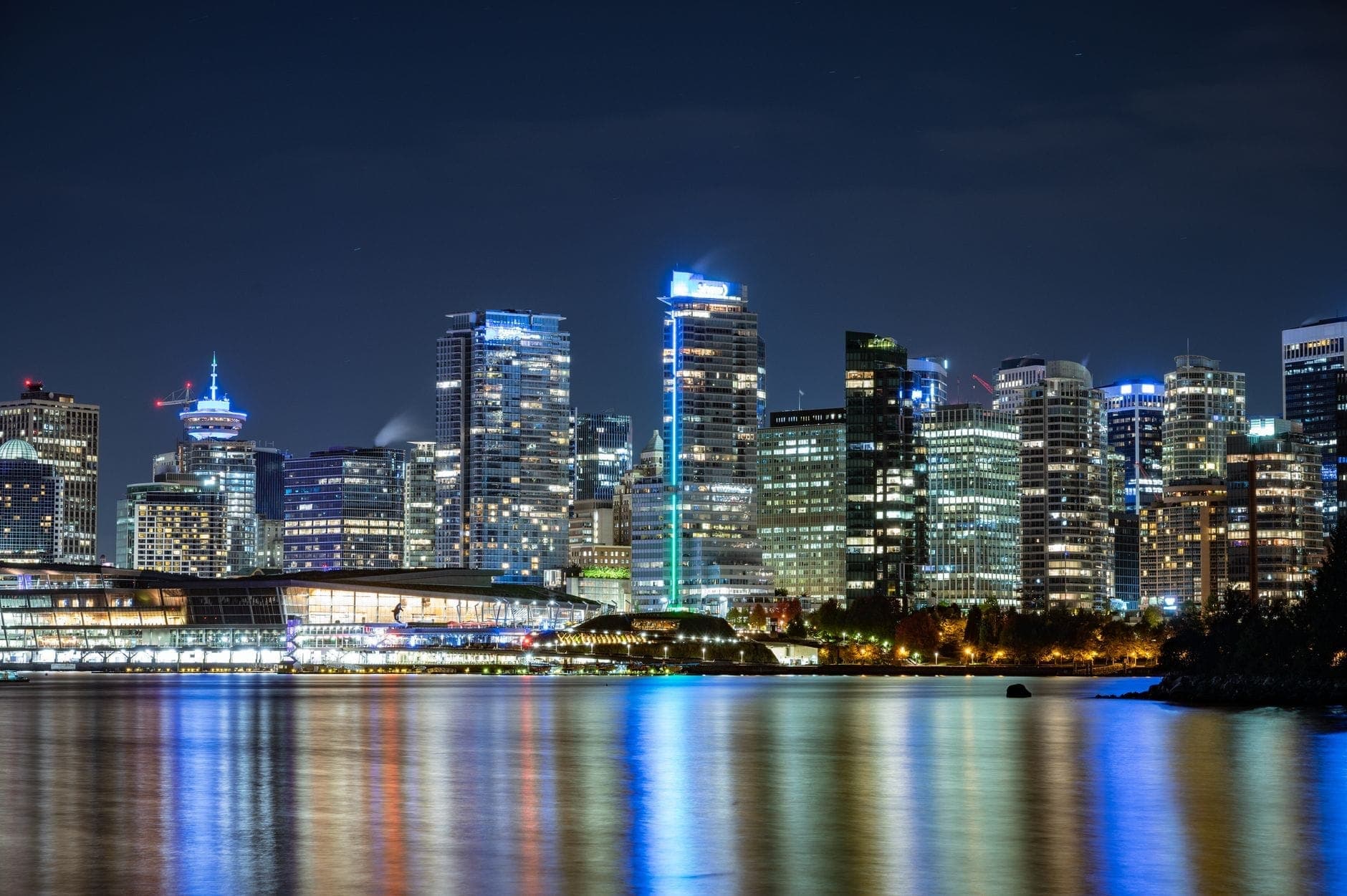 วิวยามค่ำคืนของเส้นขอบฟ้าใจกลางเมือง Vancouver พร้อมตึกระฟ้าที่สว่างไสวสะท้อนในน้ำ รวมถึงสถานที่สำคัญที่โดดเด่นและพื้นที่ริมน้ำ