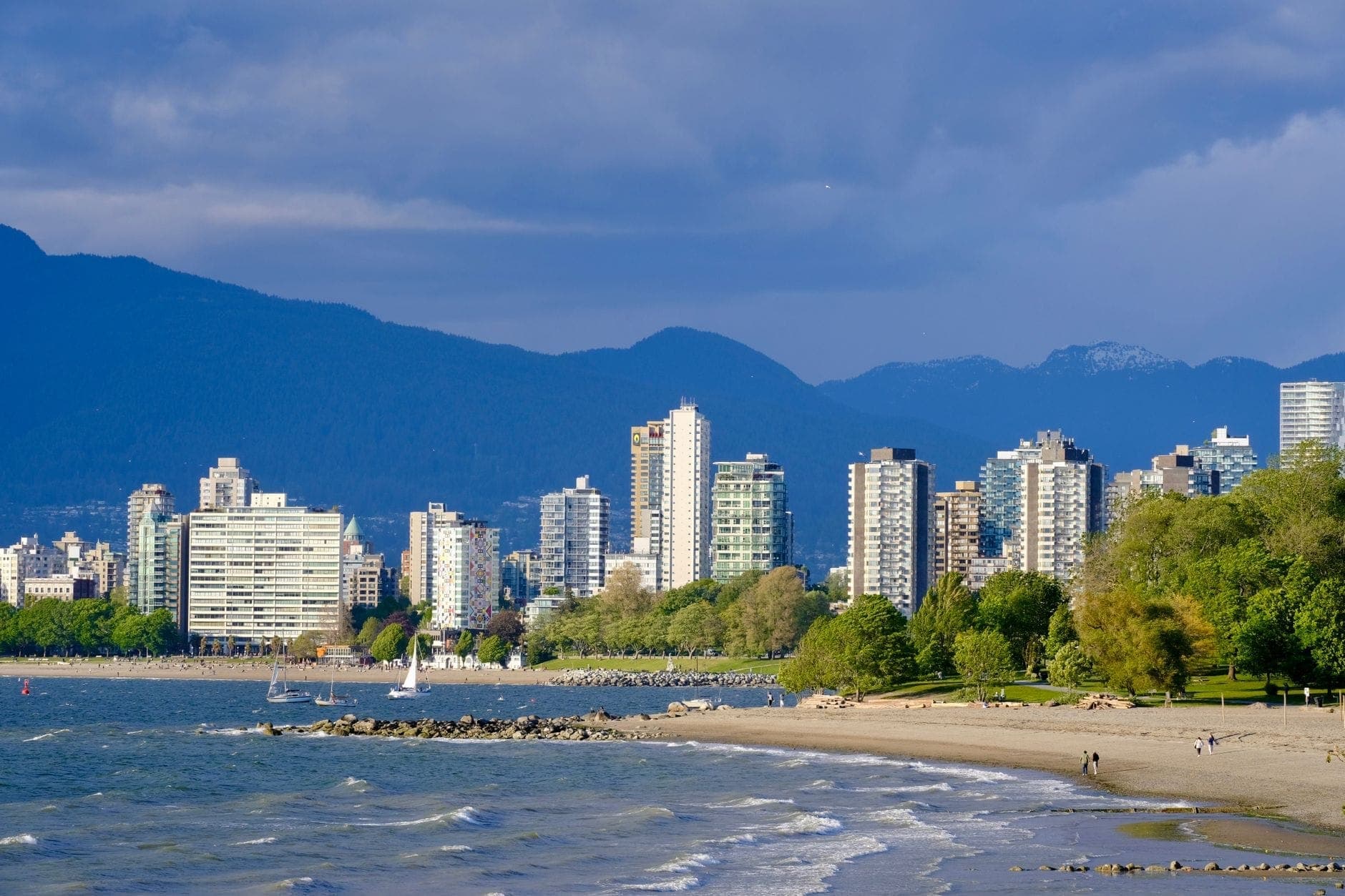 ชายหาดของ Vancouver พร้อมเส้นขอบฟ้าของเมือง ภูเขาเป็นฉากหลัง และผู้คนกำลังเดินบนทรายริมน้ำ