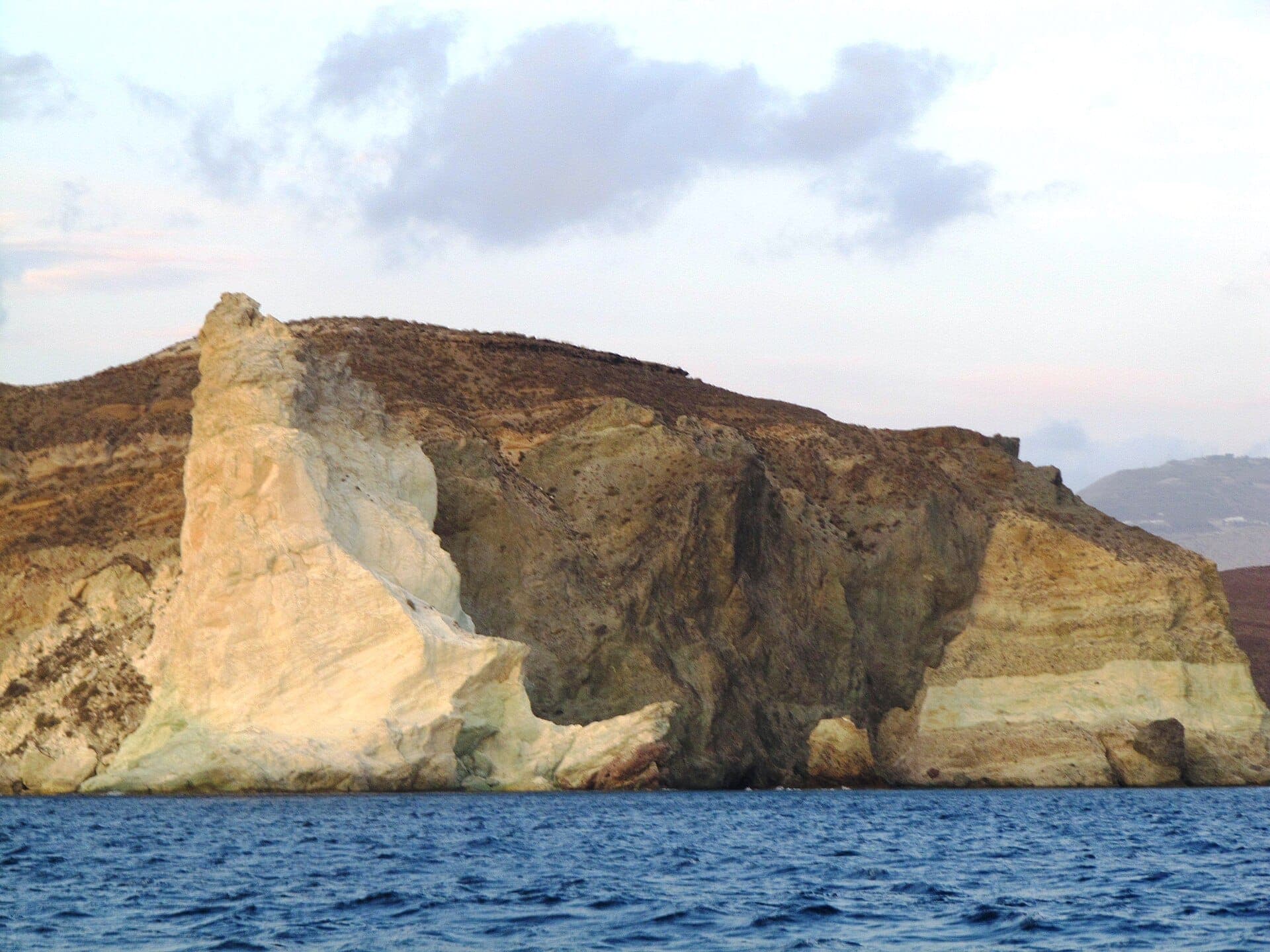 White Beach in Akrotiri, Santorini, mit aufragenden weißen Vulkanklippen, die sich über das tiefe Blau der Ägäis unter einem leicht bewölkten Himmel erheben.