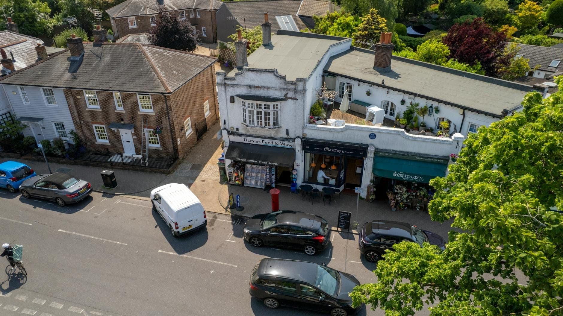 Aerial view of a pleasant London high street with small independent shops, flower stand, parked cars and tree-lined surroundings, suggesting a village-like atmosphere.