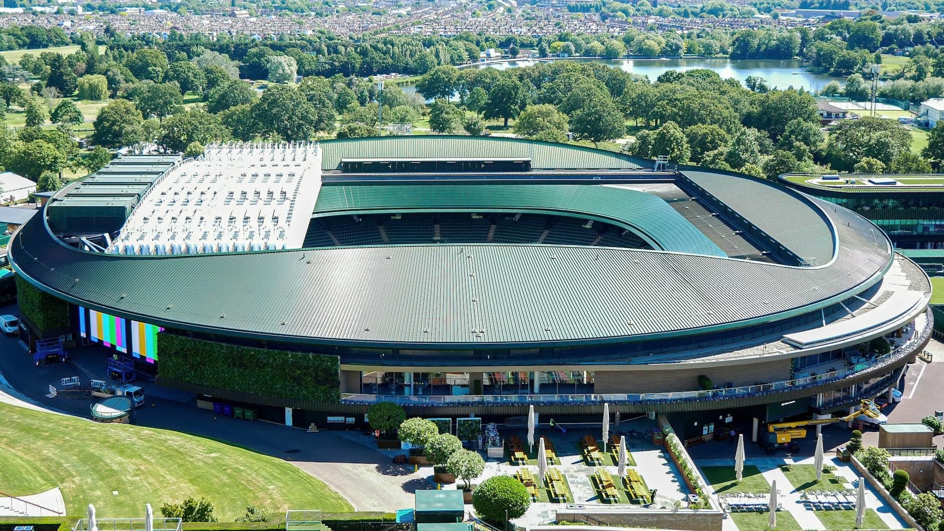 Aerial view of Wimbledon Centre Court at the All England Lawn Tennis Club with surrounding green fields and trees.