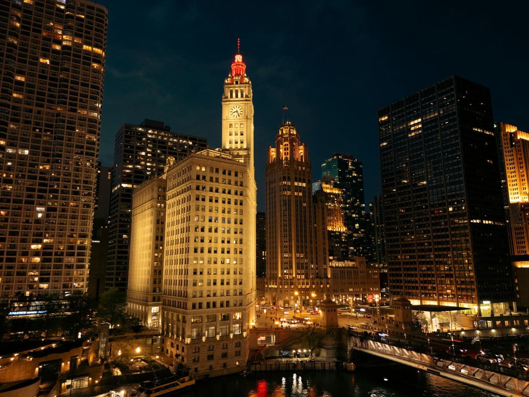 Night view of the illuminated Wrigley Building with its clock tower, surrounded by Chicago skyscrapers and the Chicago River below, creating a vibrant cityscape.