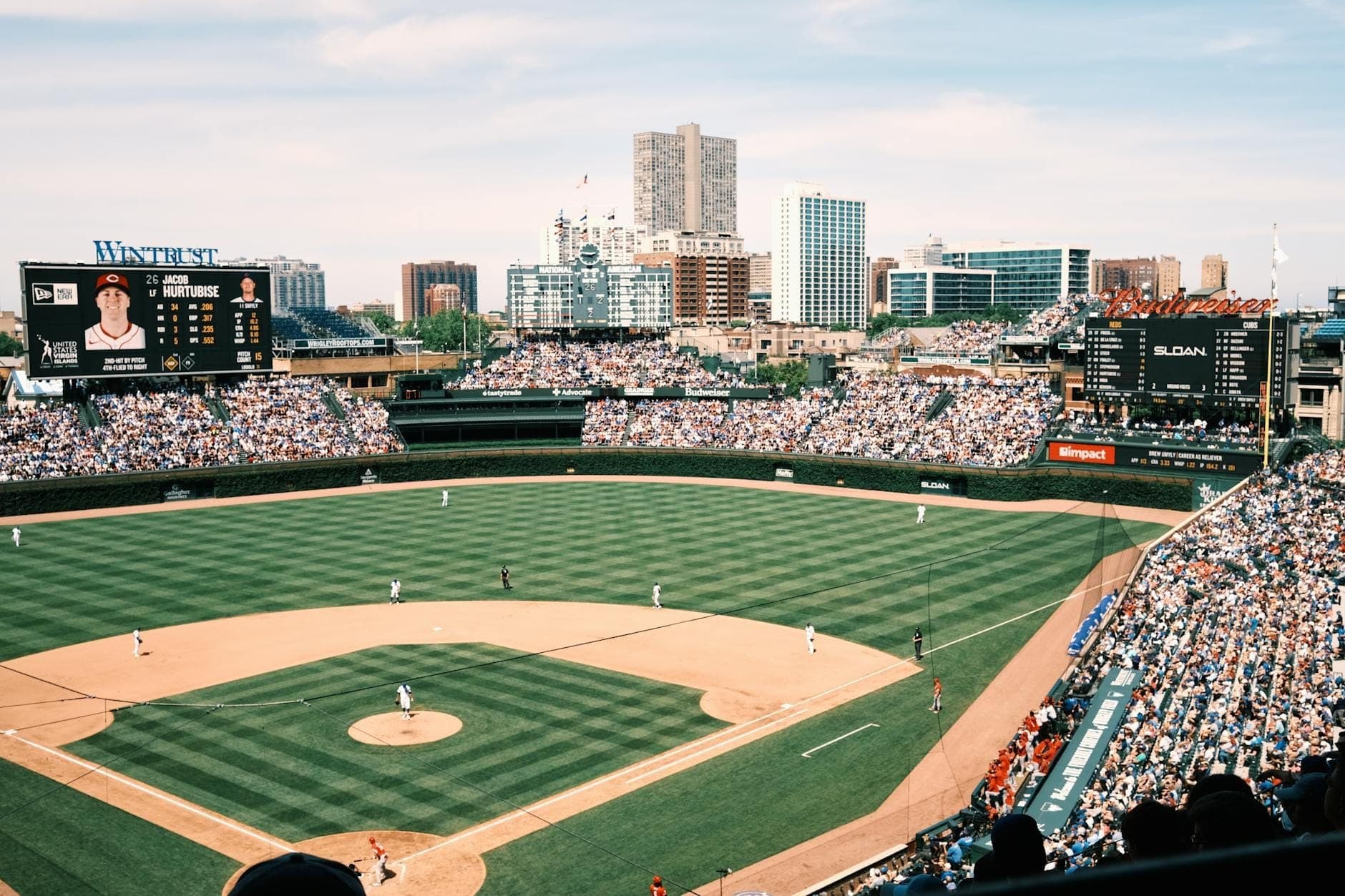 Wide landscape view of Wrigley Field during a day game, showing the green baseball diamond, packed stands, and Chicago skyline in the background.