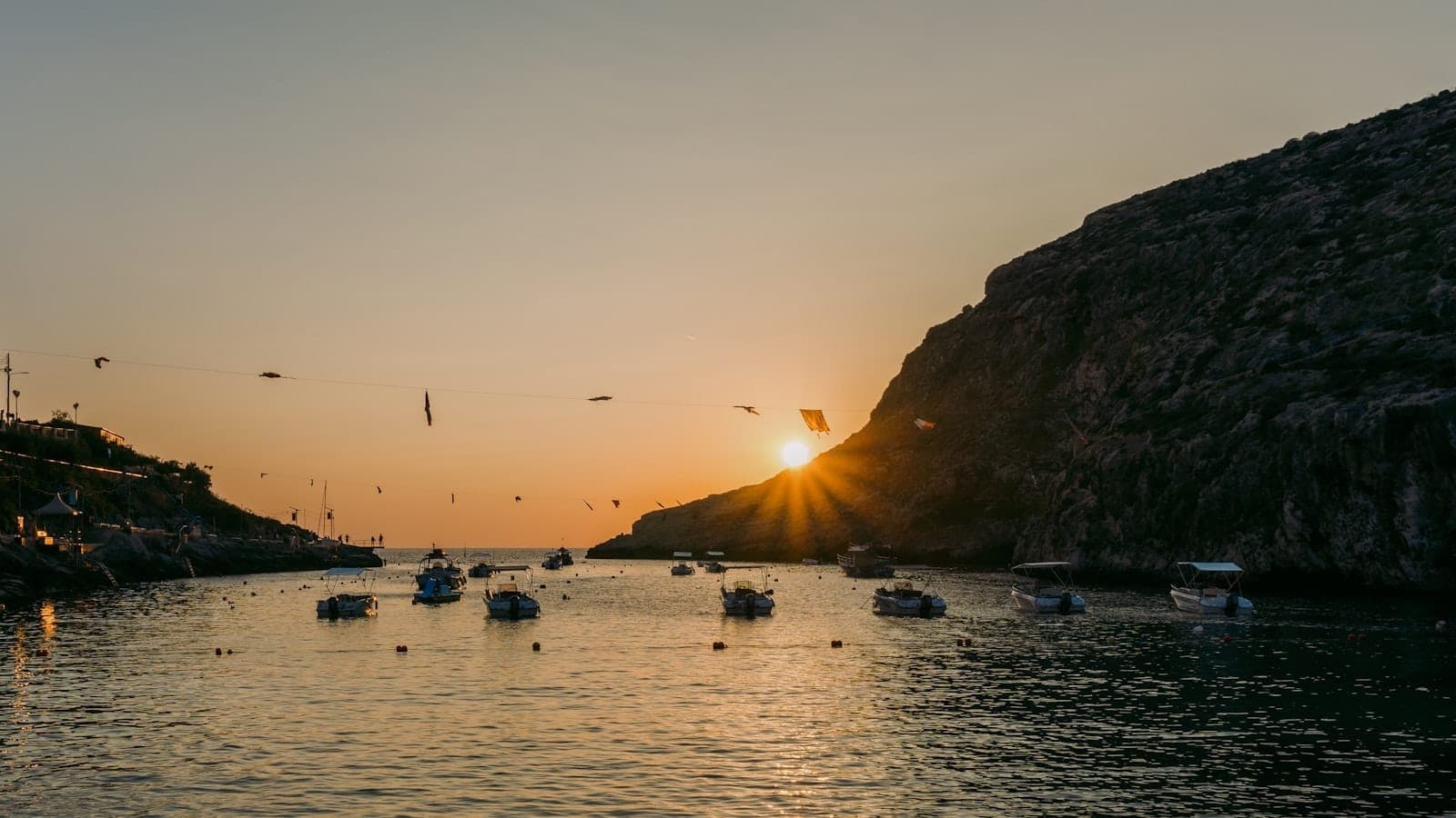 Sonnenuntergang über der Xlendi Bay mit ruhigem türkisfarbenem Wasser, kleinen Booten und dramatischen Kalksteinklippen, die die schmale Bucht einrahmen.