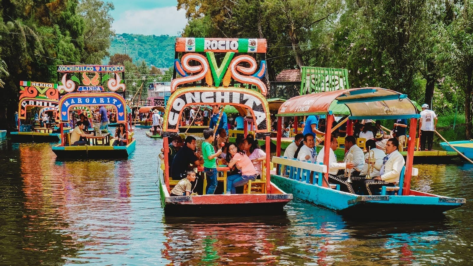 เรือตราฮิเนราสที่ทาสีสดใสเต็มไปด้วยผู้คนลอยตามคลอง Xochimilco ท่ามกลางต้นไม้สีเขียวและเงาสะท้อนบนผิวน้ำที่มีชีวิตชีวา