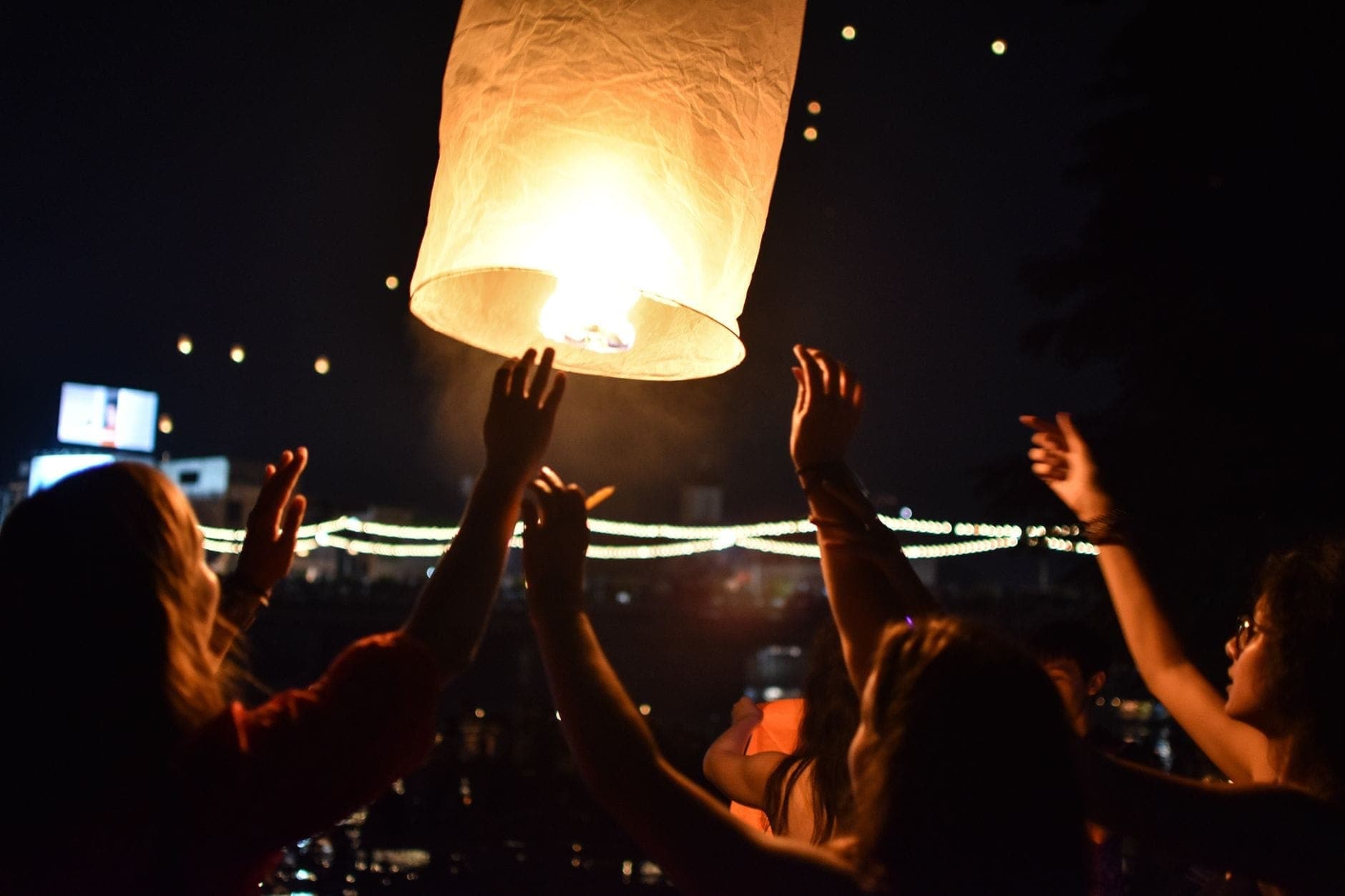 Personas soltando un farolillo luminoso por la noche durante un festival, con los brazos extendidos y el paseo fluvial iluminado al fondo.