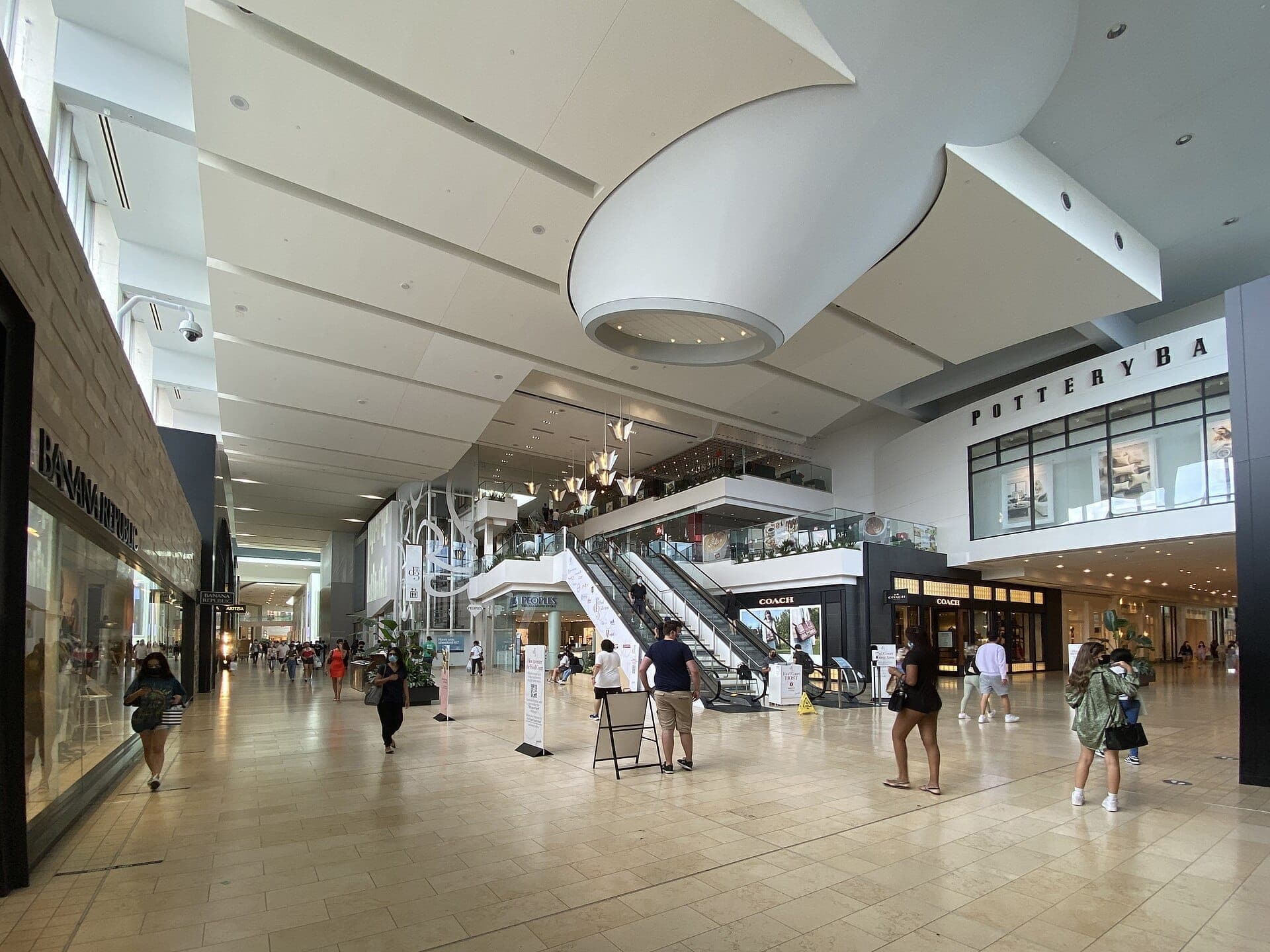 Vista interior amplia del Yorkdale Shopping Centre con arquitectura moderna, techos altos, fachadas de tiendas como Coach, Pottery Barn, y compradores caminando junto a las escaleras mecánicas.