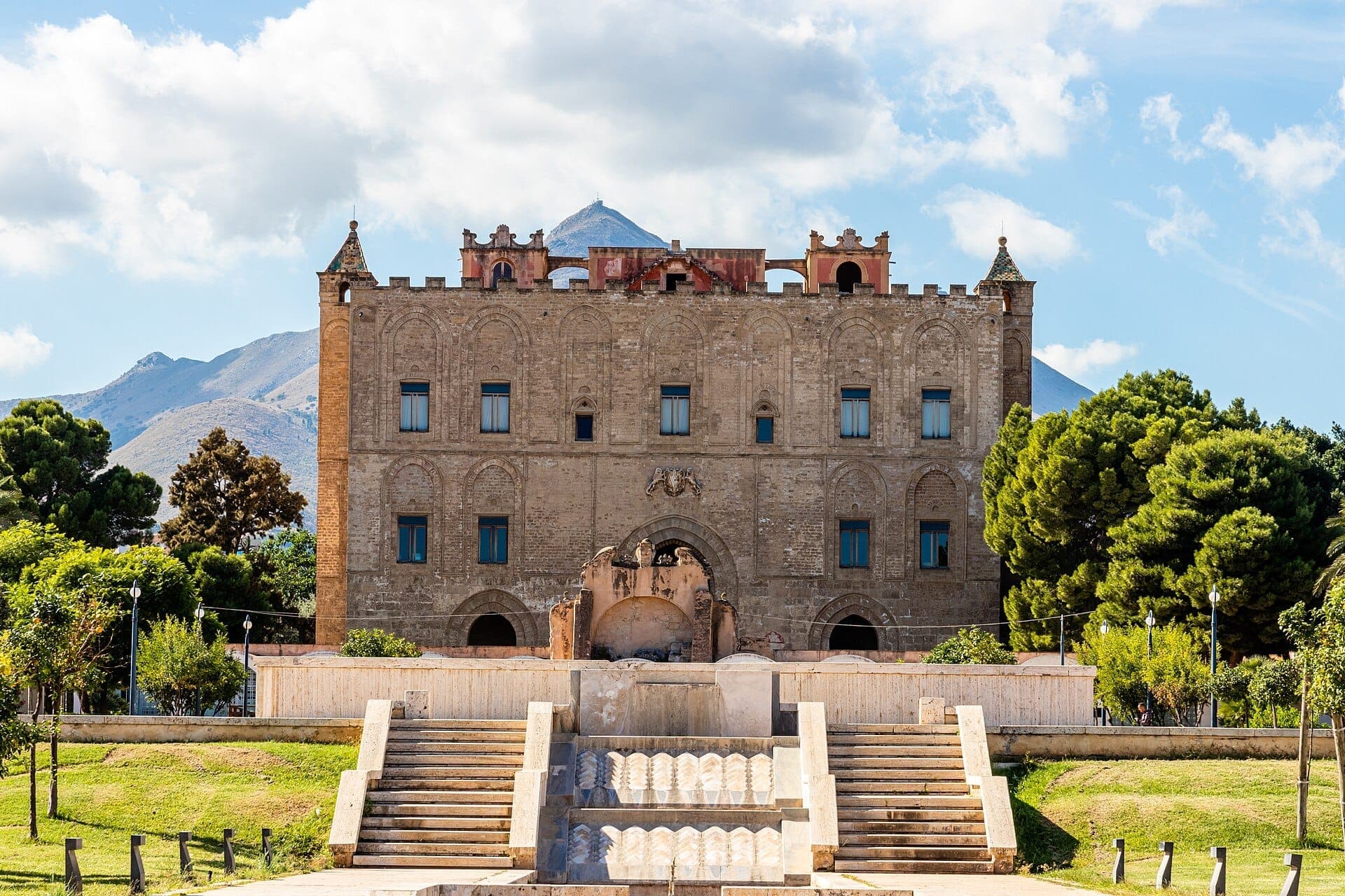 Tampak depan Kastil della Zisa di Palermo dengan arsitektur Norman yang khas, jendela melengkung besar, dan tangga batu yang dikelilingi pepohonan hijau.