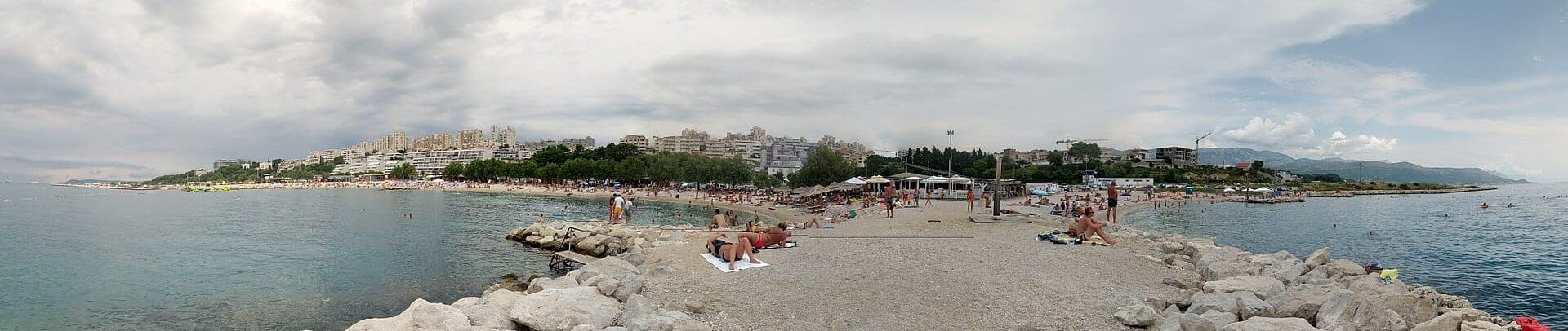 Vue panoramique de la plage de Žnjan à Split montrant le rivage de galets, des baigneurs, une eau bleue cristalline et des immeubles modernes en arrière-plan.