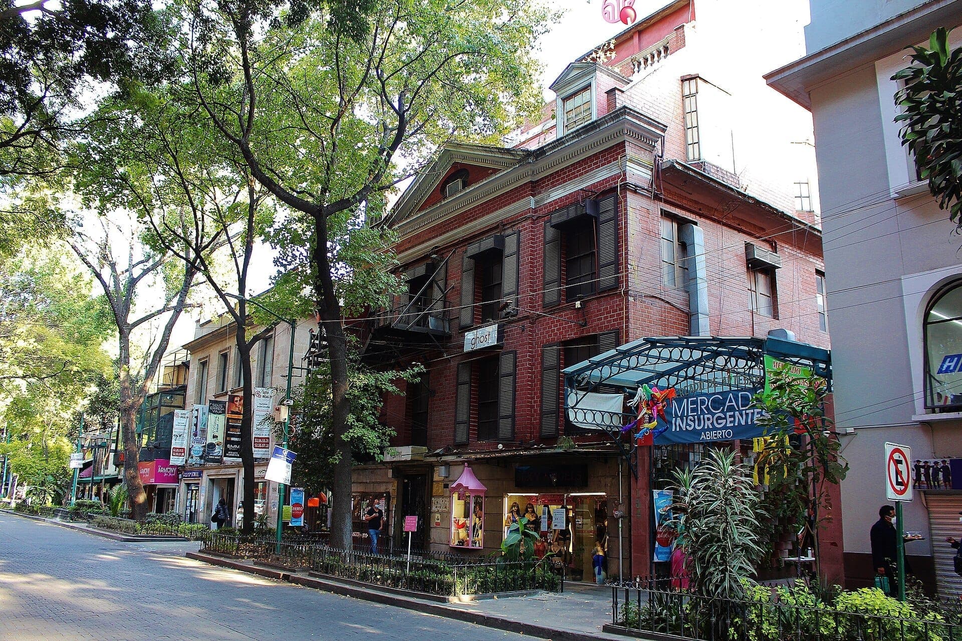 วิวถนนของ Zona Rosa ใน Mexico City ที่มีอาคารอิฐเก่าแก่ ต้นไม้ร่มรื่น และทางเข้า Mercado Insurgentes ในวันที่แดดสดใส