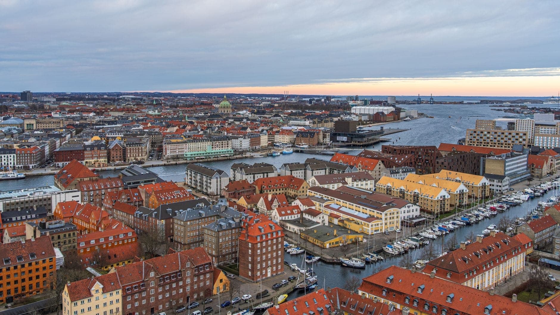 Vista aérea de Christianshavn em Copenhague mostrando edifícios coloridos à beira do canal, vias navegáveis e barcos, com a cidade e o porto ao fundo.