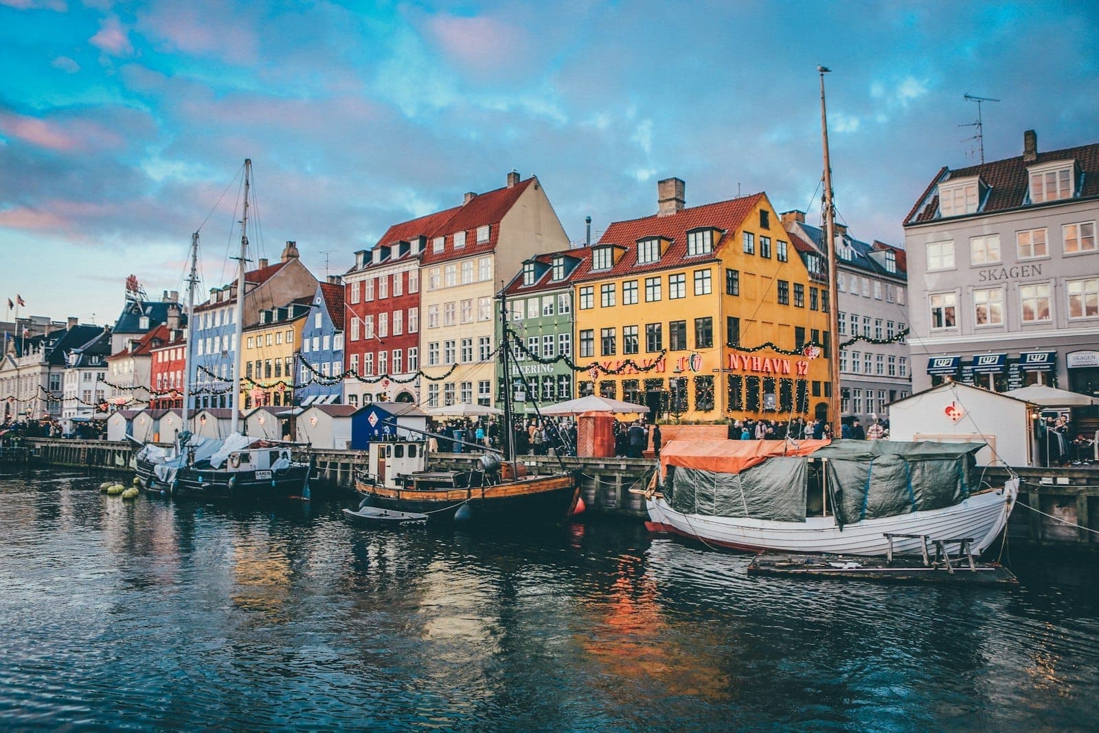 Bunte Häuser am Nyhavn-Kanal spiegeln sich bei Sonnenuntergang im Wasser, dazu festgemachte Boote und ein lebhafter Abendhimmel – das Postkartenmotiv Kopenhagens.